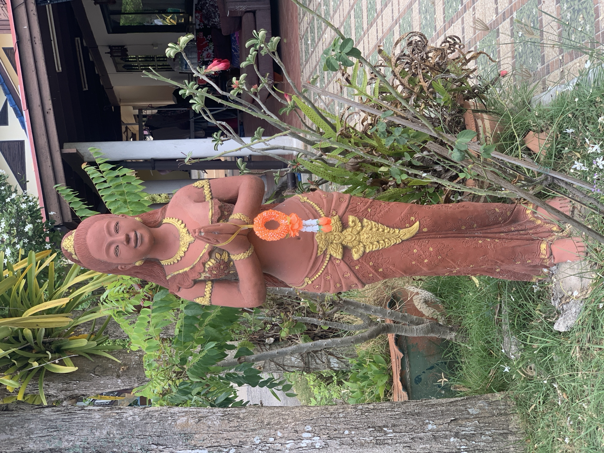 A terracotta statue of a female figure, dressed in traditional Thai attire with golden embellishments, is the focal point of the scene. She holds a garland of orange and white flowers in front of her. The statue is situated in a garden area in Baan Thong Po, Thailand. The surroundings include various plants and trees, and a building with a wooden exterior is visible in the background. The ground is covered with grass and tiles.