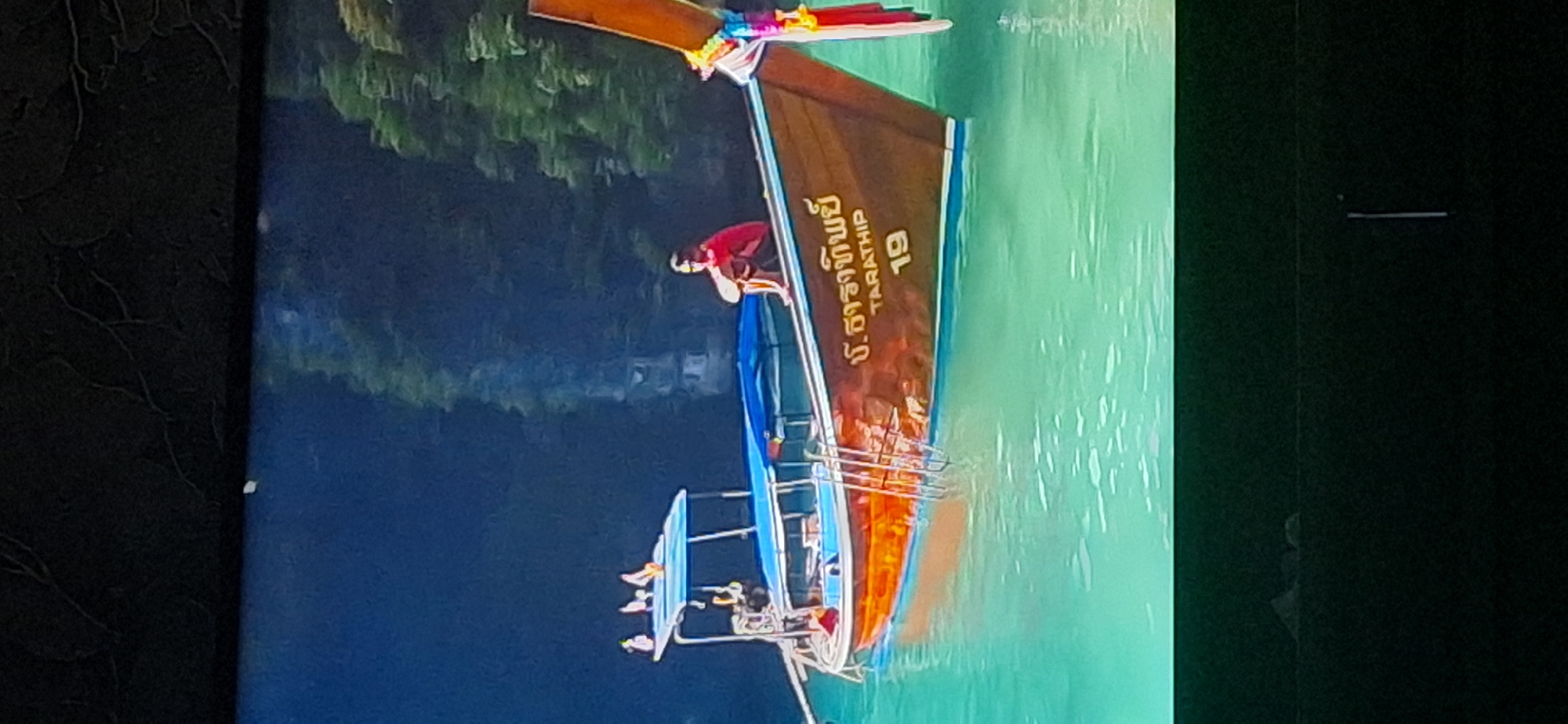 A long-tail boat is floating in turquoise water. The boat is brown with orange accents and has a blue canopy. There are ladders attached to the side of the boat. On the side of the boat, there is text in what appears to be Thai script and the words "TARATHIP" and the number "19". A person wearing a red shirt is sitting on the edge of the boat, facing away from the camera, with their legs crossed. The background consists of dark trees and foliage. The image is an interior shot of a television screen, as indicated by the dark border and the texture on the top border. There is no indication or relevance to the city of Bila Tserkva, Ukraine.