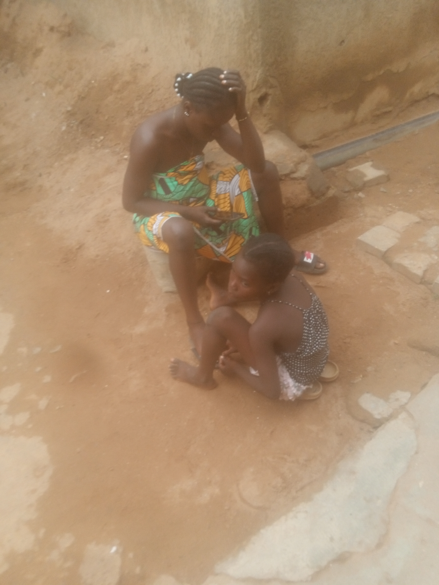 A woman and a young girl are sitting on the ground in an outdoor setting. The woman, with dark skin and braided hair adorned with small white beads, is wearing a colorful patterned fabric wrapped around her body. She has her hand on her head, suggesting a moment of reflection or perhaps discomfort. The young girl, also dark-skinned, is wearing a polka-dot patterned dress with white trim and sandals. She is sitting beside the woman, looking slightly to the left of the frame. The ground appears to be dirt or sand. In the background, there is a wall or structure. A drain pipe runs parallel along the side wall, with stones along the path of the pipe. The location is Unknown, Unknown.