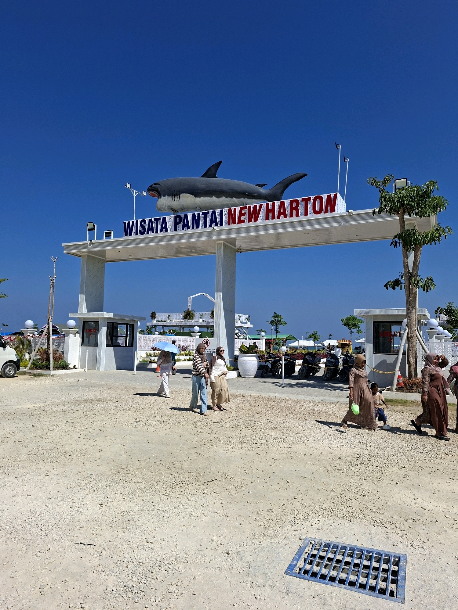 The image shows the entrance gate of "WISATA PANTAI NEW HArTON" (New Harton Beach Tour) in Camplong, Indonesia. A large, grey shark sculpture is mounted above the entrance arch. The gate is a modern, white structure with marble-like pillars. To the left of the entrance, there is a small security booth with a "LOKET" (ticket/counter) sign. Several people are walking past the entrance, some of whom are wearing traditional Indonesian clothing. In the background, motorcycles are parked and there are some trees and distant structures. The ground is covered with gravel and sand, and a blue metal grate is visible in the foreground. The sky is clear and blue.