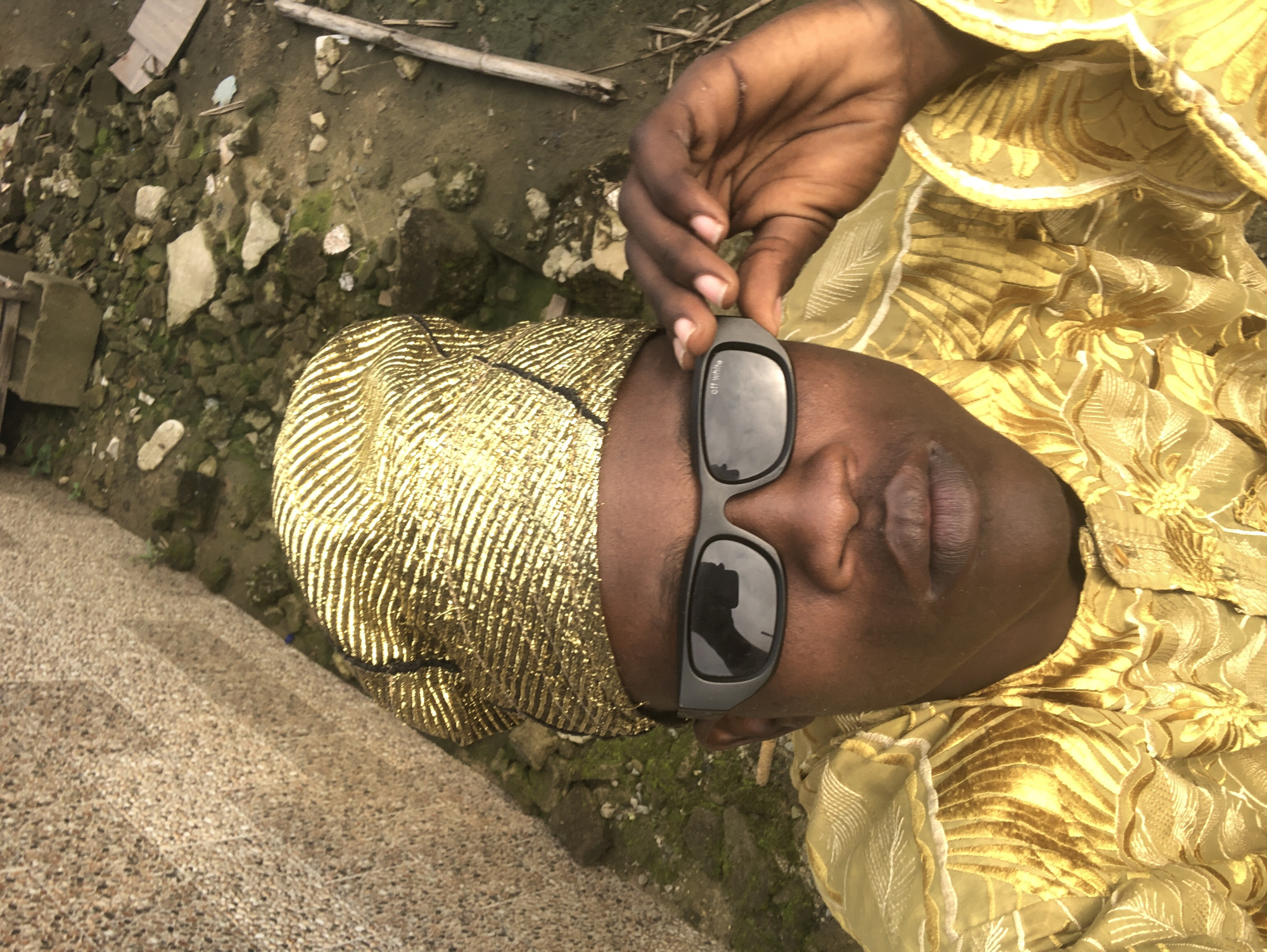 In Ado, Nigeria, a person wearing a golden turban and matching ornate attire poses for a selfie. They are adjusting dark sunglasses with "off white" text visible on the lens. The background consists of a textured wall on the left and a patch of rough, debris-strewn ground with sparse vegetation and a fallen wooden branch on the right.