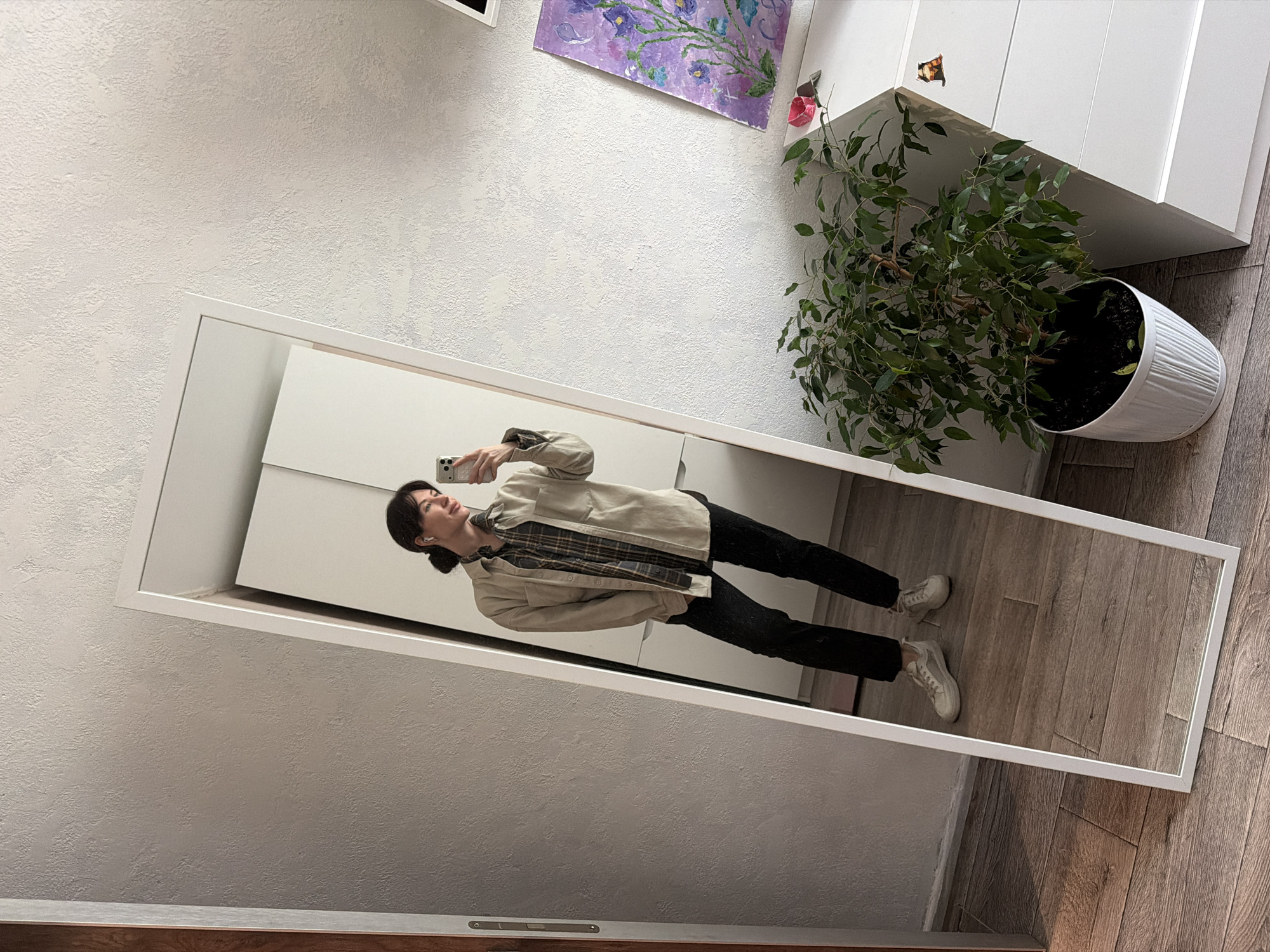 A woman with dark hair pulled back in a bun takes a mirror selfie in what appears to be an apartment in Perm, Russia. She is wearing black pants, light beige sneakers, a plaid shirt, and a light beige jacket. The mirror is framed in white and leans against a white wall, reflecting her image and a wood floor. To the right of the mirror is a white dresser with a small cabinet on top, as well as a potted plant with green leaves. On the wall above the dresser, there is a painting with purple and blue colors. The wall is textured. A dark-colored trim is visible along the bottom edge of the frame.
