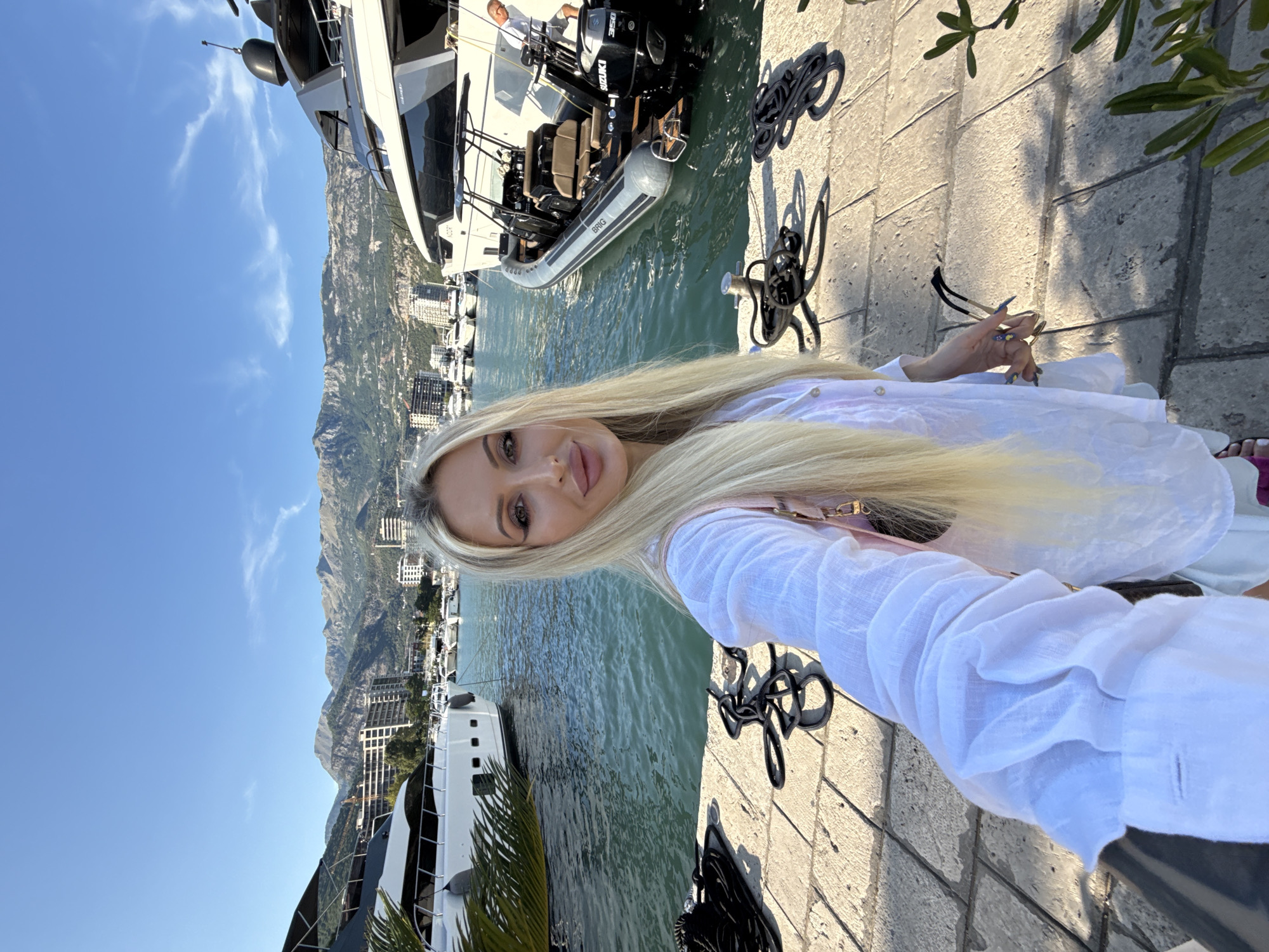 A woman with long blonde hair takes a selfie in a marina with mountains in the background. She is wearing a white linen shirt and a pink purse is slung over her shoulder. The marina is filled with yachts and boats, and the water is calm and blue. The mountains behind the marina are covered in green trees and some buildings are visible on their slopes. The sky is clear and blue with a few wispy clouds. The woman is standing on a stone walkway with ropes coiled on the ground. In the foreground, a large white yacht is docked, and a smaller inflatable boat is tied to its side. The overall scene suggests a luxurious vacation or a beautiful day out on the water. The background landscape is consistent with the Bay of Kotor, and the prominent yachts and marina structures point to Budva, Montenegro, as the location.