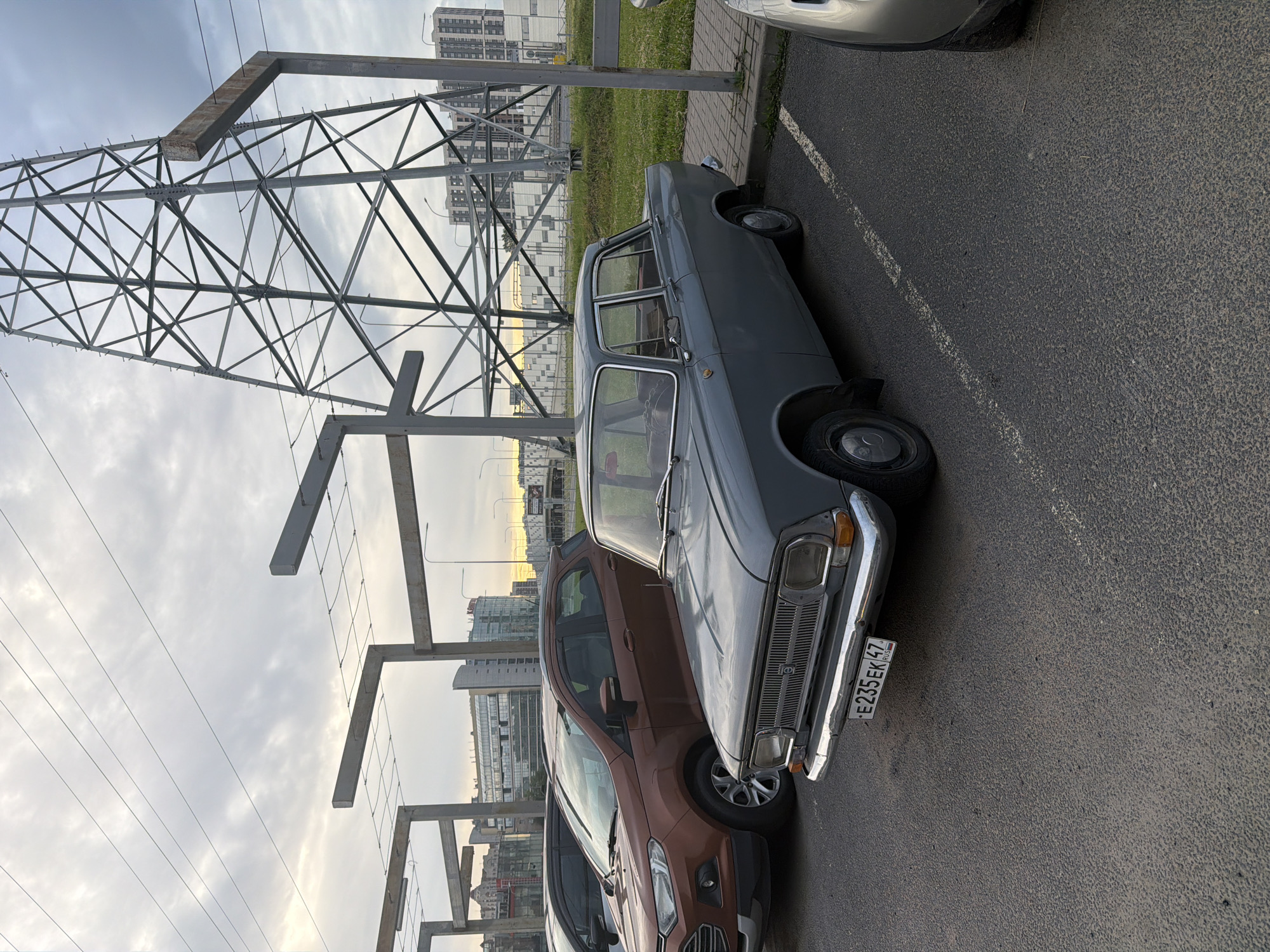 A grey, older model sedan is parked in a lot next to a brown SUV. The sedan has a license plate that reads "E235EK47 RUS". In the background, there are modern buildings and a large metal tower structure. The sky is overcast and grey.
