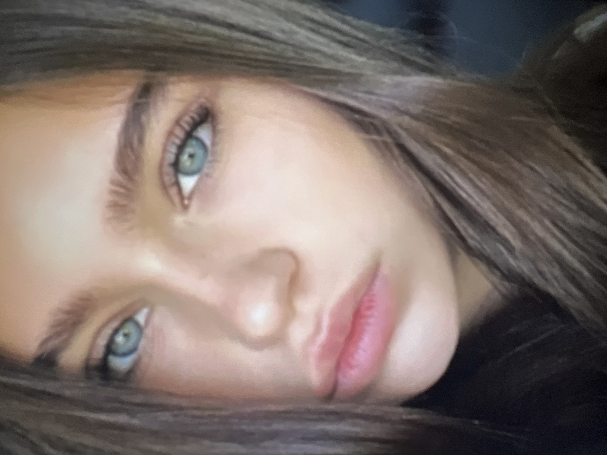 The image is a close-up, slightly blurred portrait of a young person's face, with a focus on their eyes and lips. The person has light skin, prominent eyebrows, and striking blue eyes with dark lashes. Their lips are full and appear to be naturally colored with a slight pinkish hue. Long, brown hair frames the face. The background is out of focus and dark, suggesting an indoor setting or low lighting conditions. There is no discernible information or context related to the city of Qarağandı city administration, Kazakhstan.