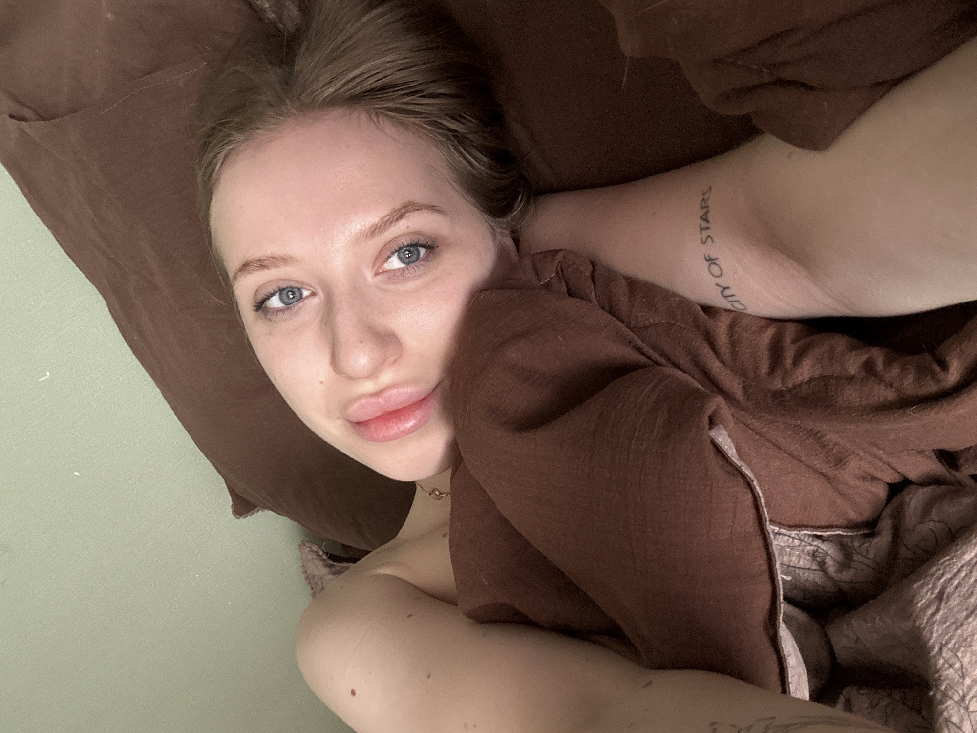 A young woman lies in bed, her head resting on a brown pillow. She has fair skin, blonde hair pulled back from her face, and blue eyes. Her lips are full and slightly parted. A brown blanket or garment is draped over her body. An arm with a tattoo reading "CITY OF STARS" is visible near her head. A wall appears to be next to the bed. The city and country are not discernible from the image.