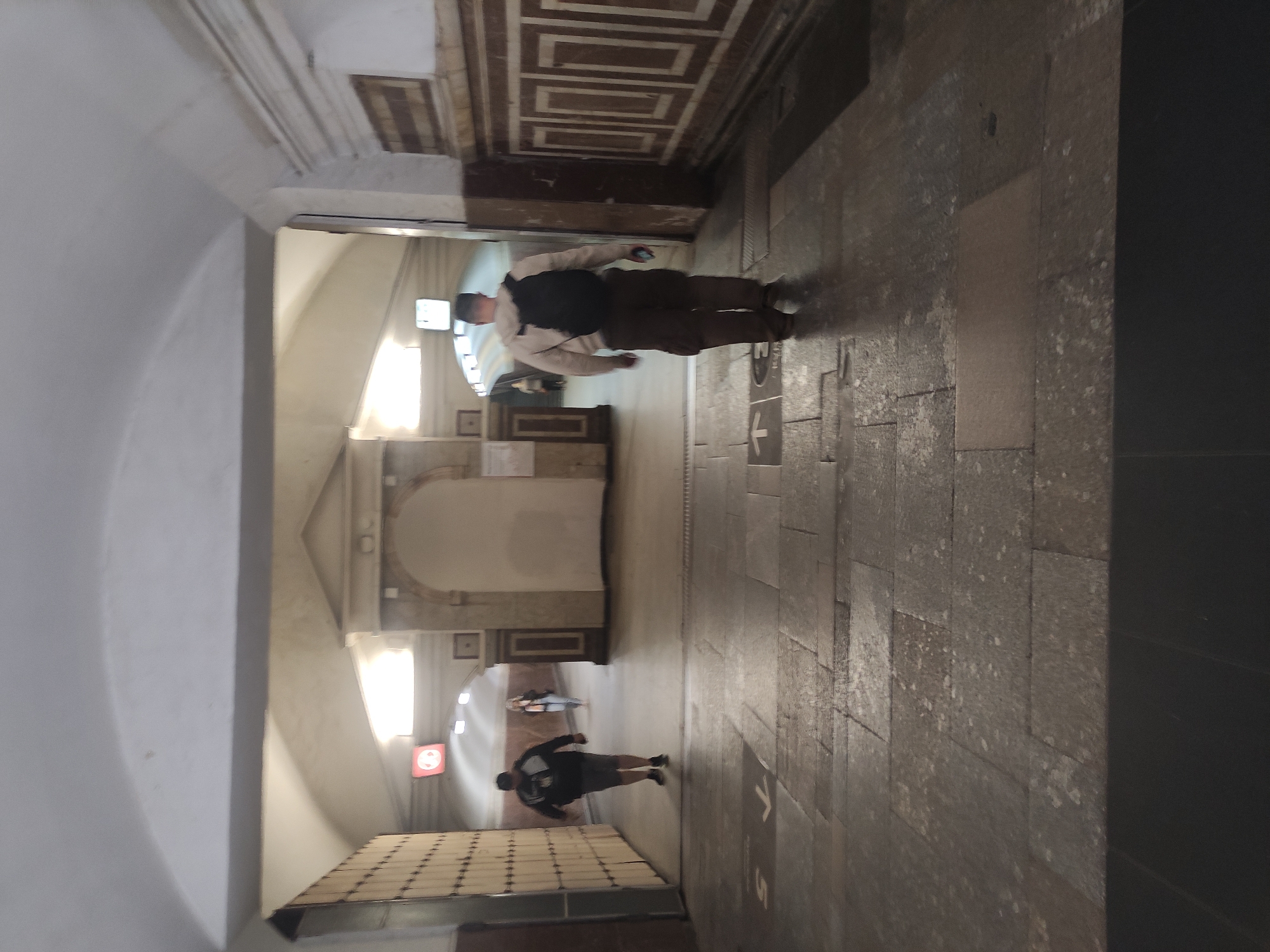 The image shows an indoor space with a person walking towards the viewer from a well-lit area. The location appears to be a corridor or passageway, characterized by stone-tiled flooring and an archway leading to another brightly lit section. On the right, a dark, smooth wall contrasts with the rough texture of the stone floor. The person walking towards the viewer is dressed in casual clothing. In the distance, there are additional people and architectural details suggesting a public space, such as a museum or historical building. The presence of directional arrows on the floor indicates the path of travel.