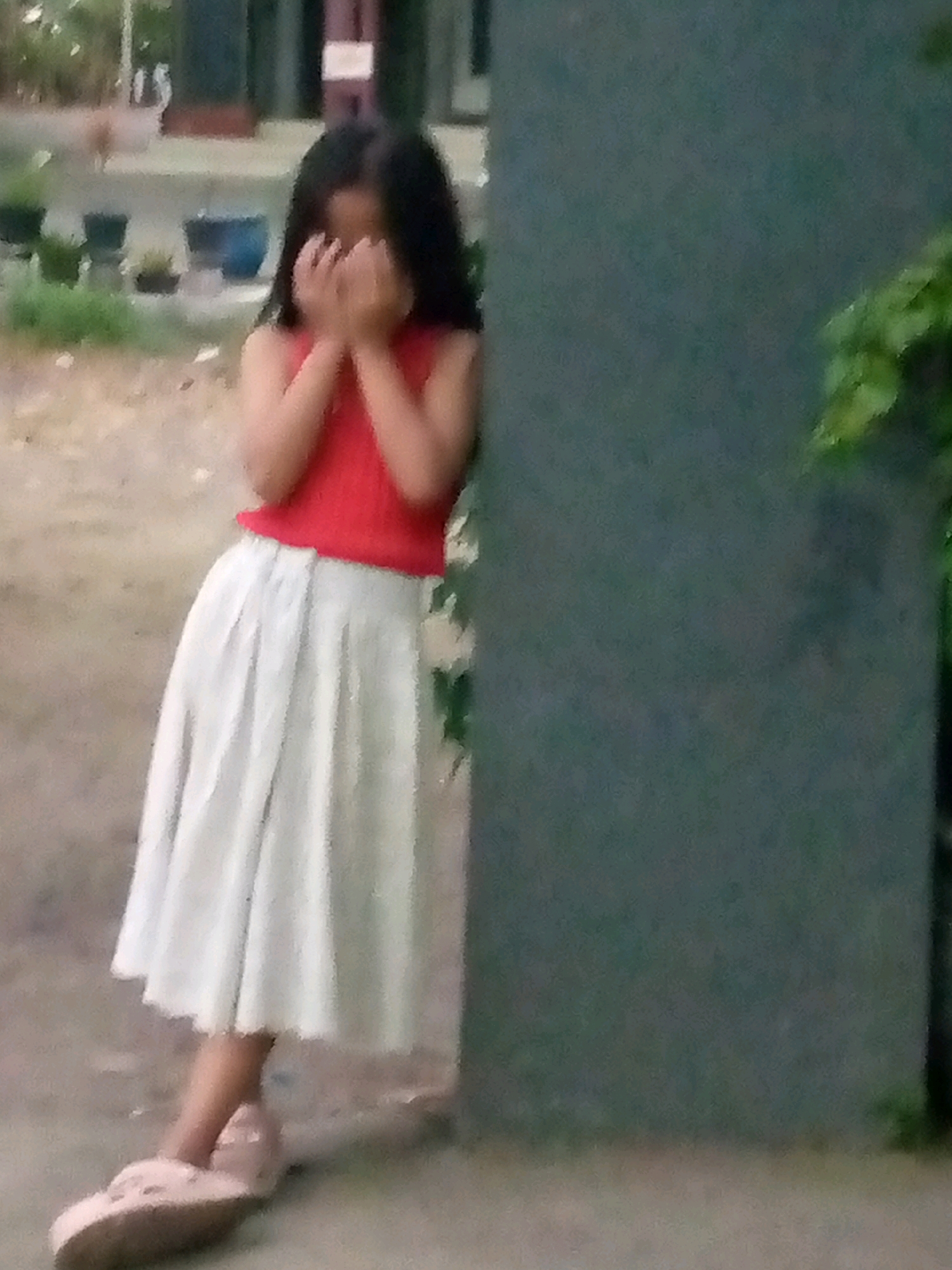 In a street scene in Sumberwono, Indonesia, a young girl with dark hair stands leaning against a dark green textured wall. She is wearing a red sleeveless top and a long, light-colored pleated skirt. Her hands are covering her face, suggesting she might be shy or distressed. Her feet are clad in light pink slip-on sandals. The background shows a blurred outdoor environment with some vegetation and what appears to be buildings or structures. The ground beneath her is a mix of dirt and possibly gravel. The overall image quality is somewhat grainy and out of focus.