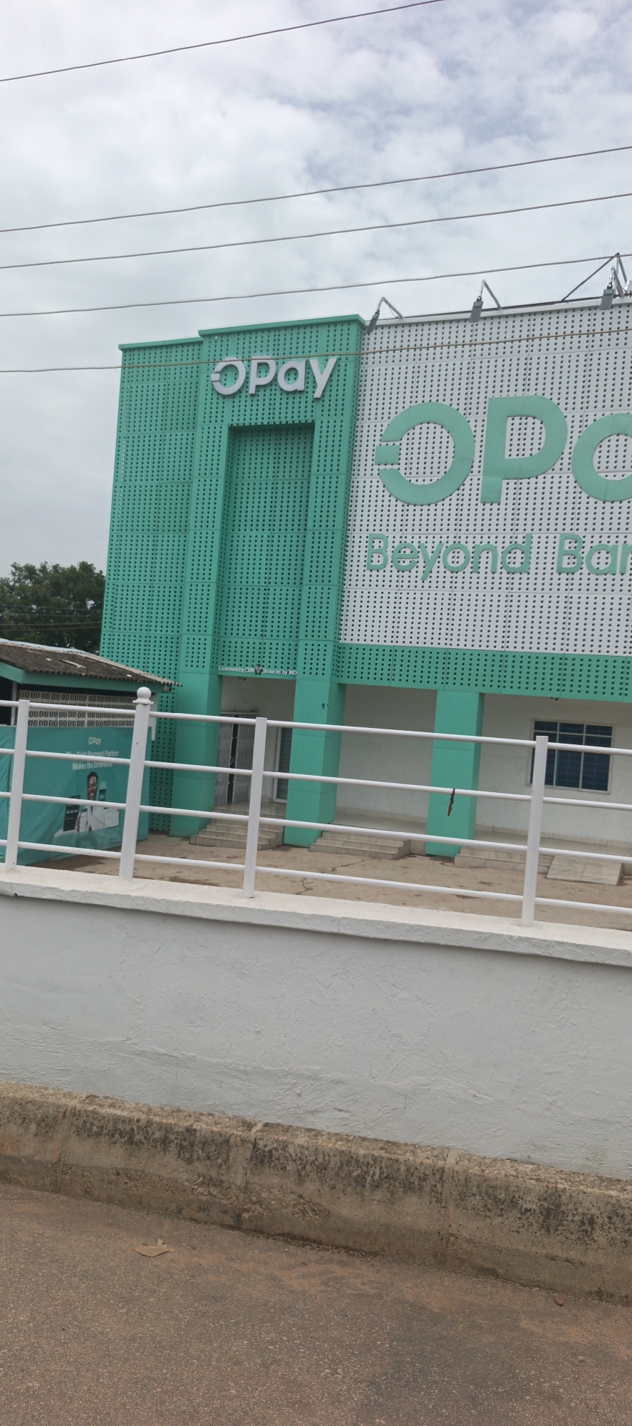 The image shows an OPay branch in Jos, Nigeria. The building is two-toned with the left side in a turquoise perforated pattern and the right side in white with turquoise OPay logo and "Beyond Banking" text, also in turquoise. A white railing runs along a white wall in front of the building. The ground is asphalt. Overhead power lines are visible. The sky is overcast.