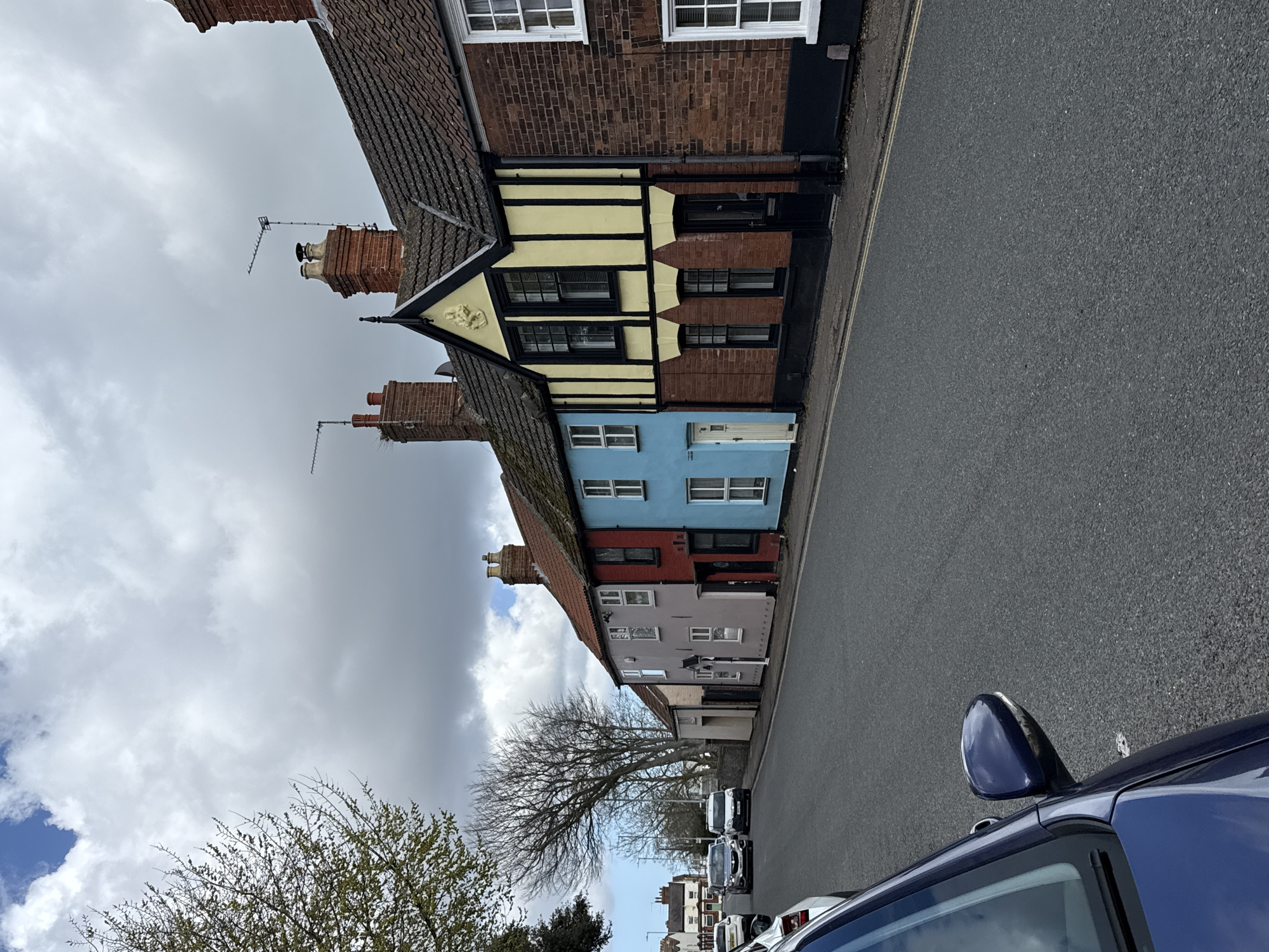 The image depicts a street in Gorleston-on-Sea, United Kingdom, featuring a row of terraced houses with varied colorful facades. The houses, mostly two-story, display colors like red, blue, pink, brown, and yellow with black and white Tudor-style elements. Chimneys and TV aerials are visible on the roofs of some houses. The road in front of the houses appears gray and asphalt. Several parked cars line the street on the far side of the road. A blue car is prominently positioned on the right edge of the frame, capturing part of its side mirror. Trees with bare branches are visible in the foreground, suggesting it might be autumn or winter. The sky is overcast with gray clouds. No people are visible.