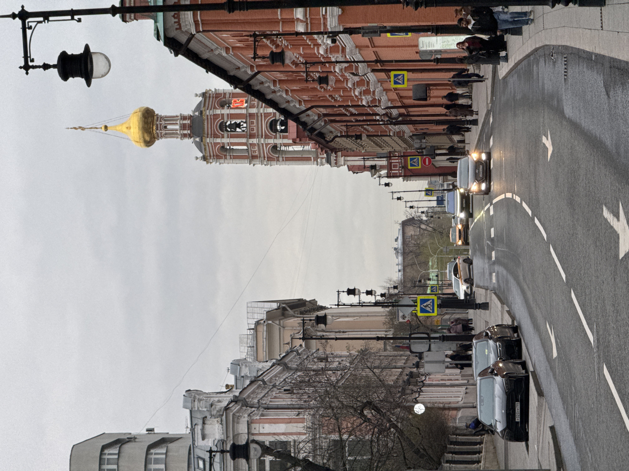 The image depicts a street scene in Moscow, Russia, featuring a view of the Petrovka Street leading towards the High-Petrov Monastery. The street is lined with buildings of varying architectural styles, including the distinct Russian Orthodox architecture of the monastery with its golden dome. People are walking along the sidewalks on both sides of the street. Several cars are visible on the wet asphalt road, moving in one direction. The sky is overcast. Street lamps are visible along the street, casting minimal light on the scene. Pedestrian crossing signs are posted along the sidewalks.