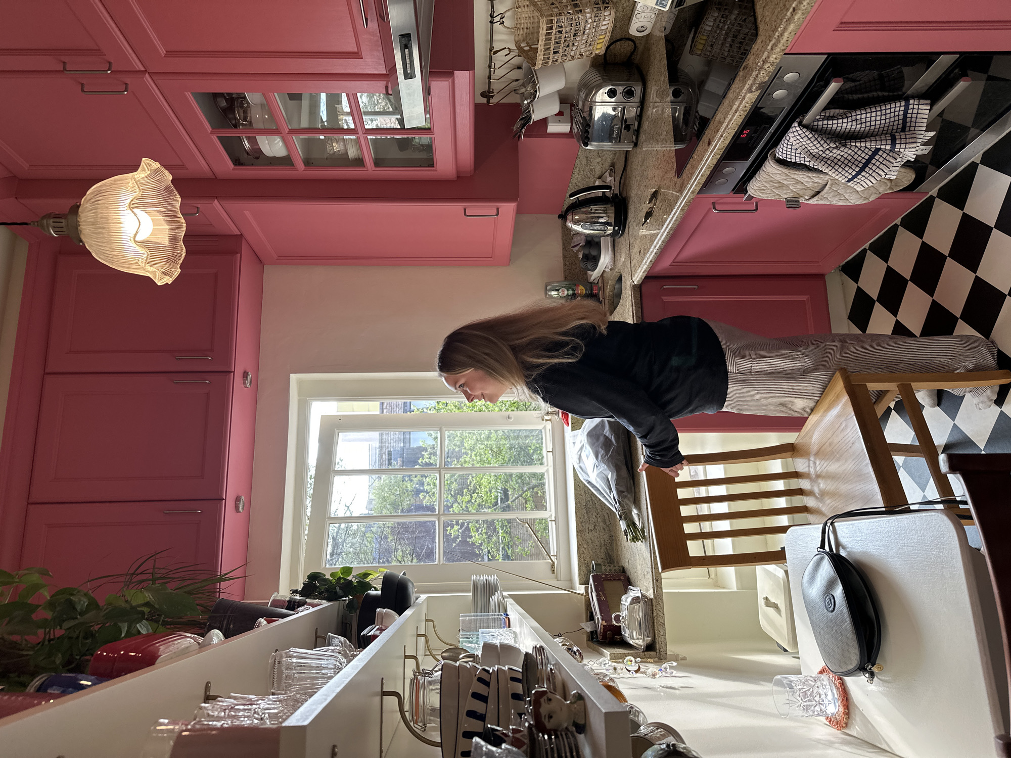 The image depicts the interior of a kitchen with pink cabinetry and a black and white checkered floor, giving the viewer an off-balance, disoriented perspective. A woman with long blonde hair is shown bending forward towards the sink area. Through a window is visible greenery and the cityscape suggestive of an urban environment. The presence of housewares such as dishes and silverware in the drying rack area near the sink further indicates the space is a kitchen. The city of London and the United Kingdom are not discernible from the provided imagery.