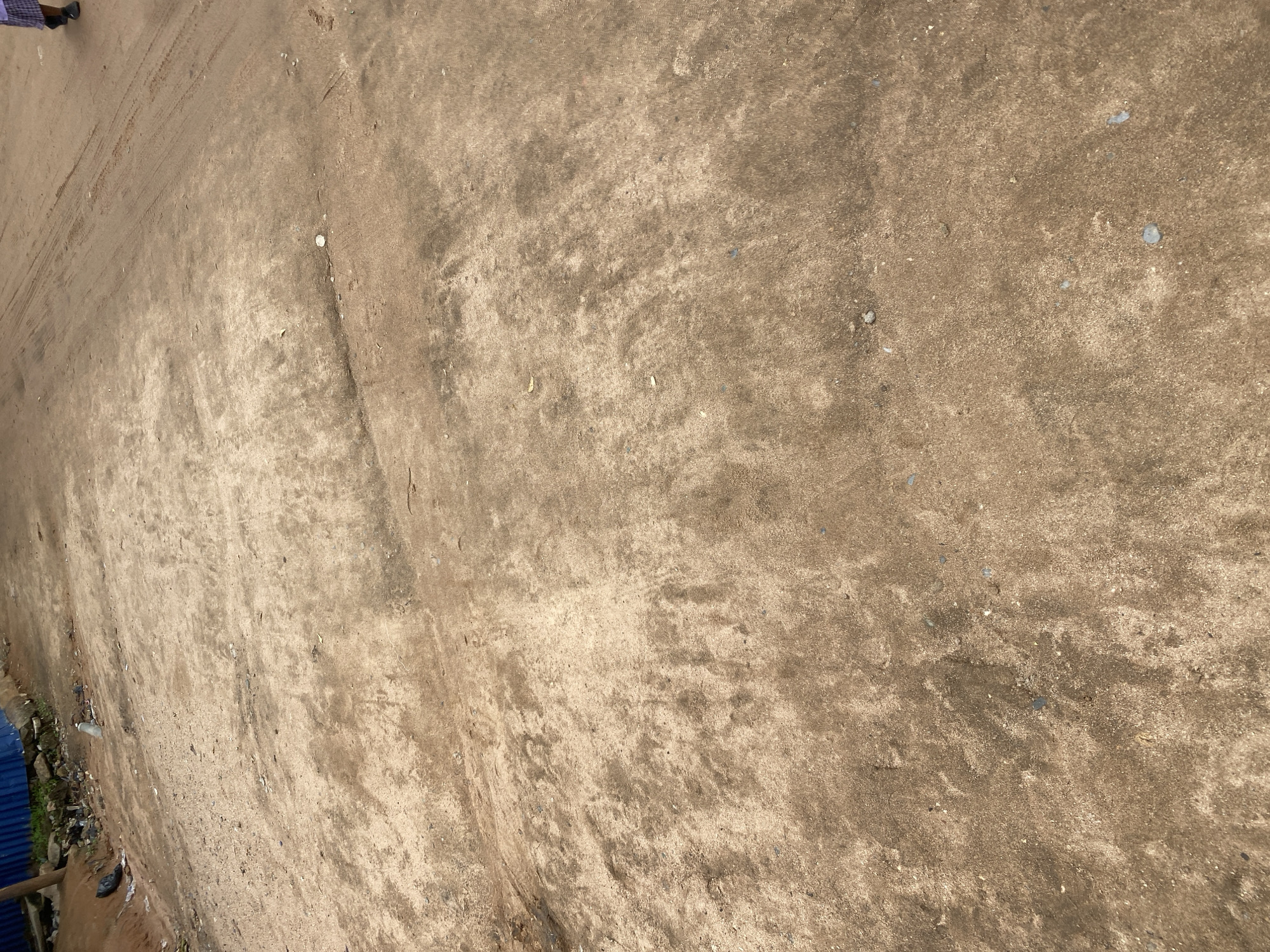 This media file shows a top-down view of a dusty, unpaved ground surface. The ground appears to be dirt or sand, with visible tire tracks and some small pebbles or debris scattered across it. The overall color is a light brown, with darker patches indicating dampness or different soil composition.
In the upper left corner, a person's legs and feet are visible, wearing dark-colored shoes and light-colored pants or shorts. The person is walking on the dusty ground, leaving faint footprints. To the far left edge, a blue corrugated metal structure is partially visible, suggesting it might be a fence or part of a building. Some discarded plastic trash and possibly some green vegetation are also visible along the edge of this structure.
The scene appears to be outdoors in daylight, as indicated by the lighting and shadows, although the exact time of day is not definitively determinable. The weather seems dry, given the dusty nature of the ground. There are no explicit cues about the specific emotions being conveyed, but the scene suggests an ordinary, possibly informal, outdoor setting in Jalingo, Nigeria. There is no visible text in the image.
