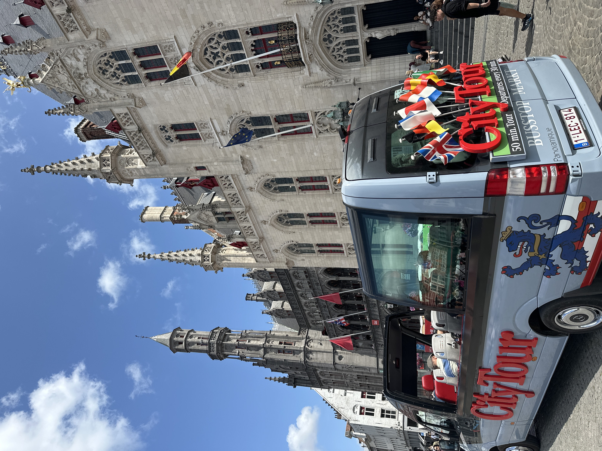 The image depicts a light blue open-top tour van, branded "City Tour" in large red lettering, parked on a cobblestone street in Bruges, Belgium. The van is adorned with numerous small flags of various countries, protruding from its rear. Behind the van, a grand Gothic-style building with intricate stone carvings and arched windows stands prominently under a bright blue sky with scattered clouds. This building appears to be the Belfry of Bruges, or a similar historical landmark in the city center. Several people are visible on the steps leading up to the building and walking on the street. The van has "50 min. Tour," "Departures: every 30 min," and "BUSSTOP MARKT" printed on its rear. The license plate of the van reads "1-LEC-814."