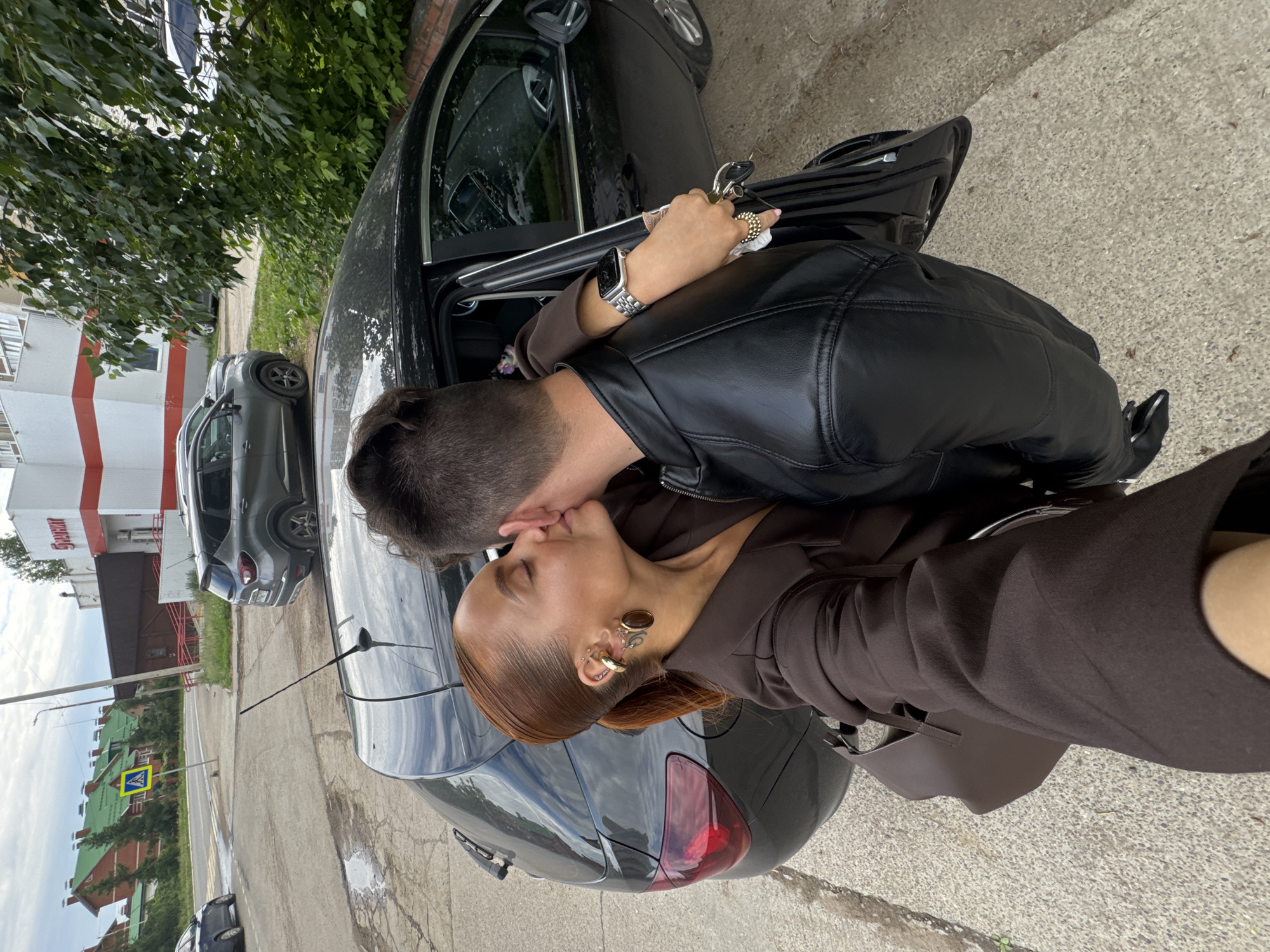 The image depicts two individuals, a man and a woman, kissing next to an open black hatchback vehicle on a paved surface in the city of Chaikovsky, Russia.
The woman, on the left, has reddish-brown hair styled into a ponytail. She is wearing a dark brown long-sleeved top and has a brown handbag over her left shoulder. Her left ear features multiple gold-colored piercings: two hoops, a large cuff, several small studs, and a prominent oval-shaped earring. She is kissing the man's left ear/cheek.
The man, with his back mostly to the camera, is on the right. He is wearing a black leather jacket with visible stitching on the shoulders. His left arm is around the woman's shoulder, and his left hand holds car keys and a small white item. He wears a silver-colored smartwatch with a matching band on his left wrist.
The central vehicle is a black hatchback, possibly a Nissan Note, with its front passenger door open. The asphalt pavement beneath them shows cracks and a small puddle.
In the background, to the right, is a white building with prominent red vertical stripes, bearing a partial "ПЯТЁРОЧКА" (Pyaterochka) sign, indicating a supermarket. Behind it, a brown building with a red-railed access ramp is visible. Further in the background, residential buildings with red brick facades and distinctive green sloped roofs are present. A blue and white pedestrian crossing sign on a leaning pole is visible on the left side of the street. Other vehicles include a dark grey SUV parked on the right and a dark blue sedan partially visible on the far left. The sky is overcast. Green foliage is present on the right side of the scene.