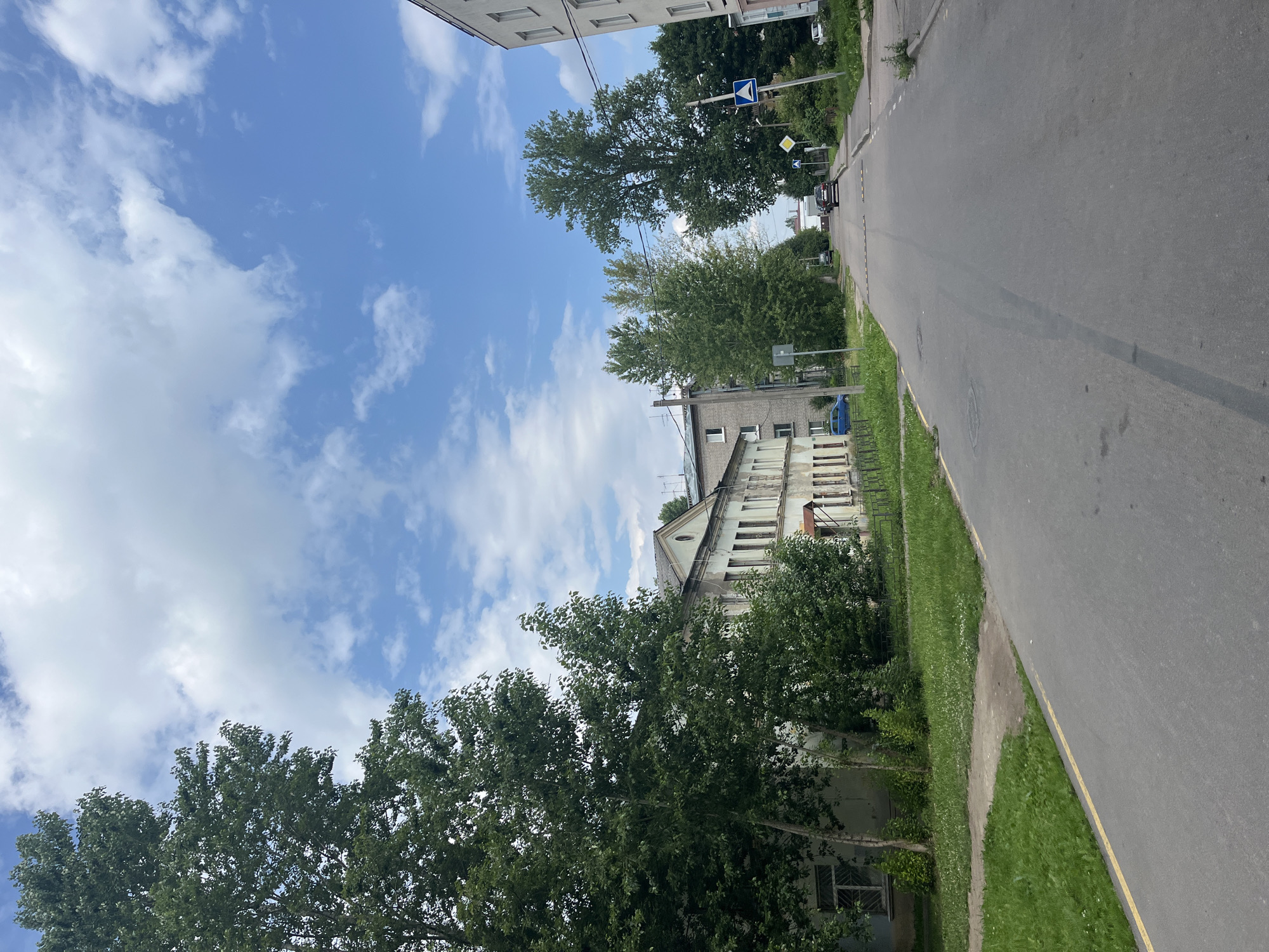 The image shows a residential street in the city of Imeni Sverdlova, Russia, under a partly cloudy sky. On the left, a large, leafy green tree partially obscures a light-colored building with multiple windows. Further down the street, more trees line the road. Several other buildings are visible, including a taller, modern-looking structure on the right. Cars are parked and driving on the asphalt road, which has yellow and white markings. Speed bumps are present on the road. The overall environment appears to be a quiet urban setting during the daytime.