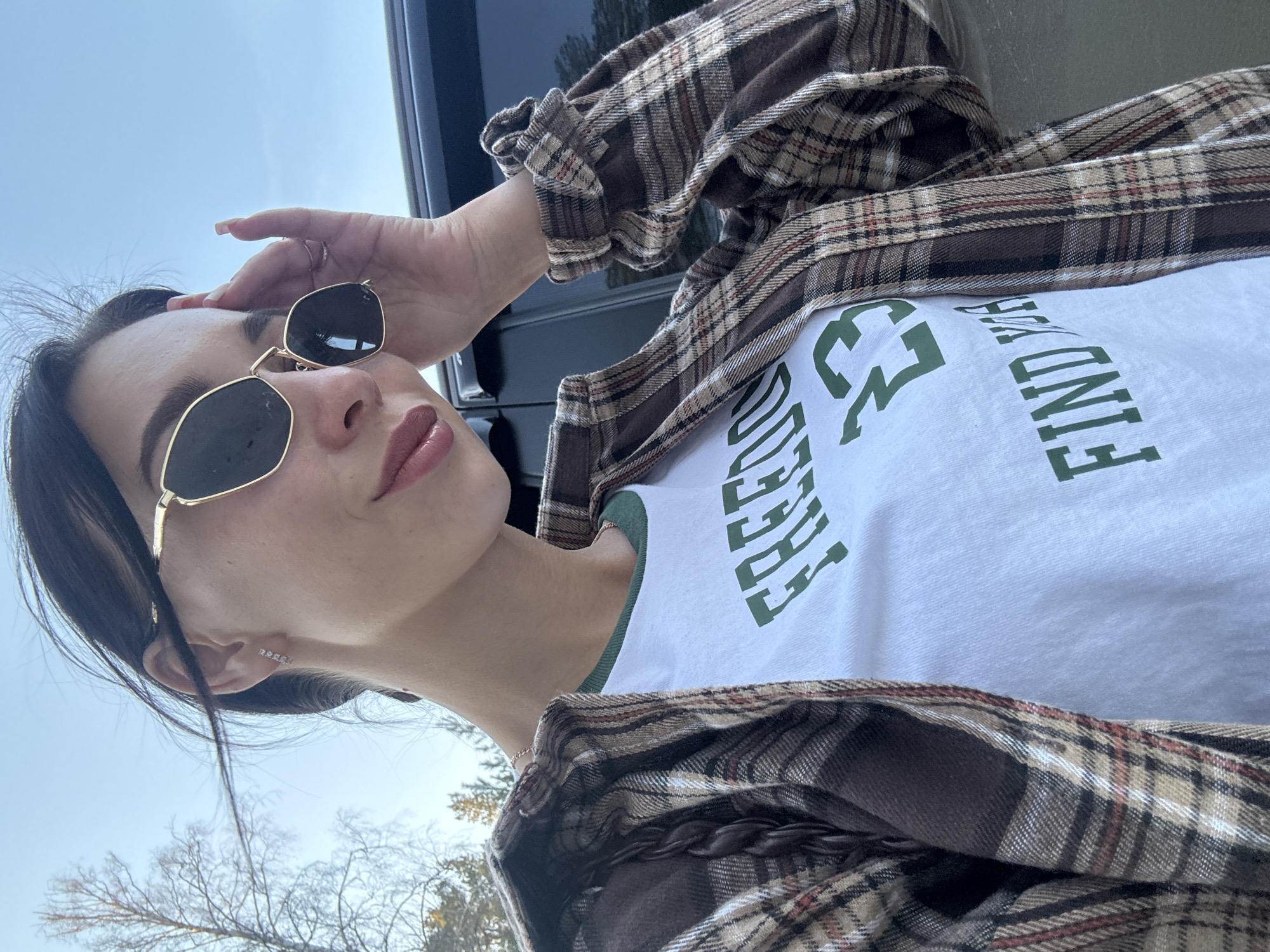 A young woman wearing a brown and tan plaid button-down shirt over a white t-shirt with green lettering poses for a selfie outdoors. She is wearing gold-rimmed sunglasses and an earring. Her dark hair is pulled back. She has one hand on her forehead, partially obscuring her face. Behind her is a black vehicle and trees. The sky is visible in the background. The location is not identifiable as Noviy, Russia, based on the visual information provided.