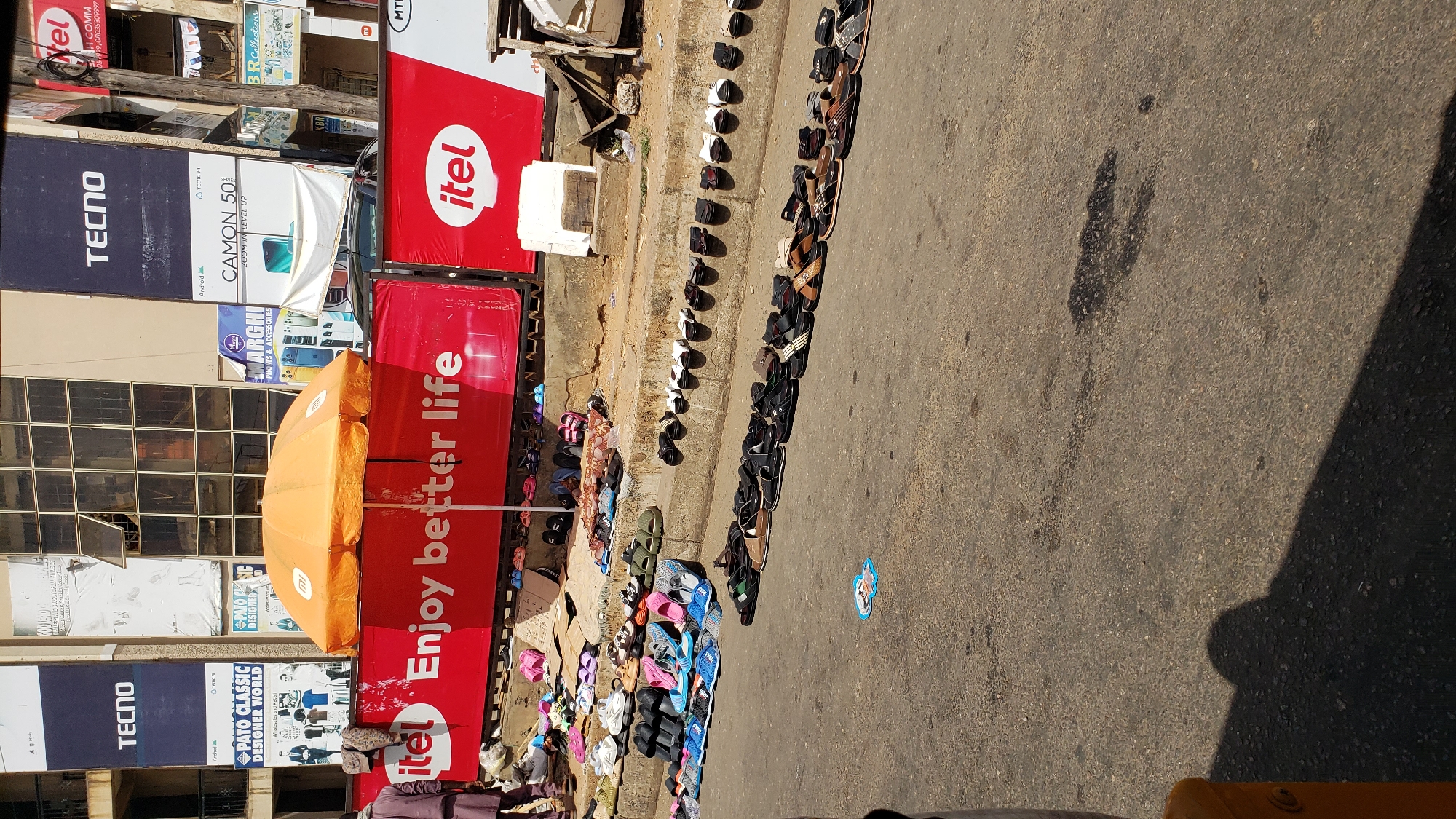 A ground-level shot depicts a street scene in Maiburiji, Nigeria. The image is oriented with the buildings and signage visible to the left. Prominent signs for "TECNO" and "itel" are stacked vertically along the side of a building. An orange umbrella stands in front of a red sign with the phrase "Enjoy better life." A large assortment of sandals and shoes is lined up along the curb of the street, suggesting a vendor selling footwear. There are no people clearly visible. A sticker rests on the road near the shoes. The street is made of asphalt.