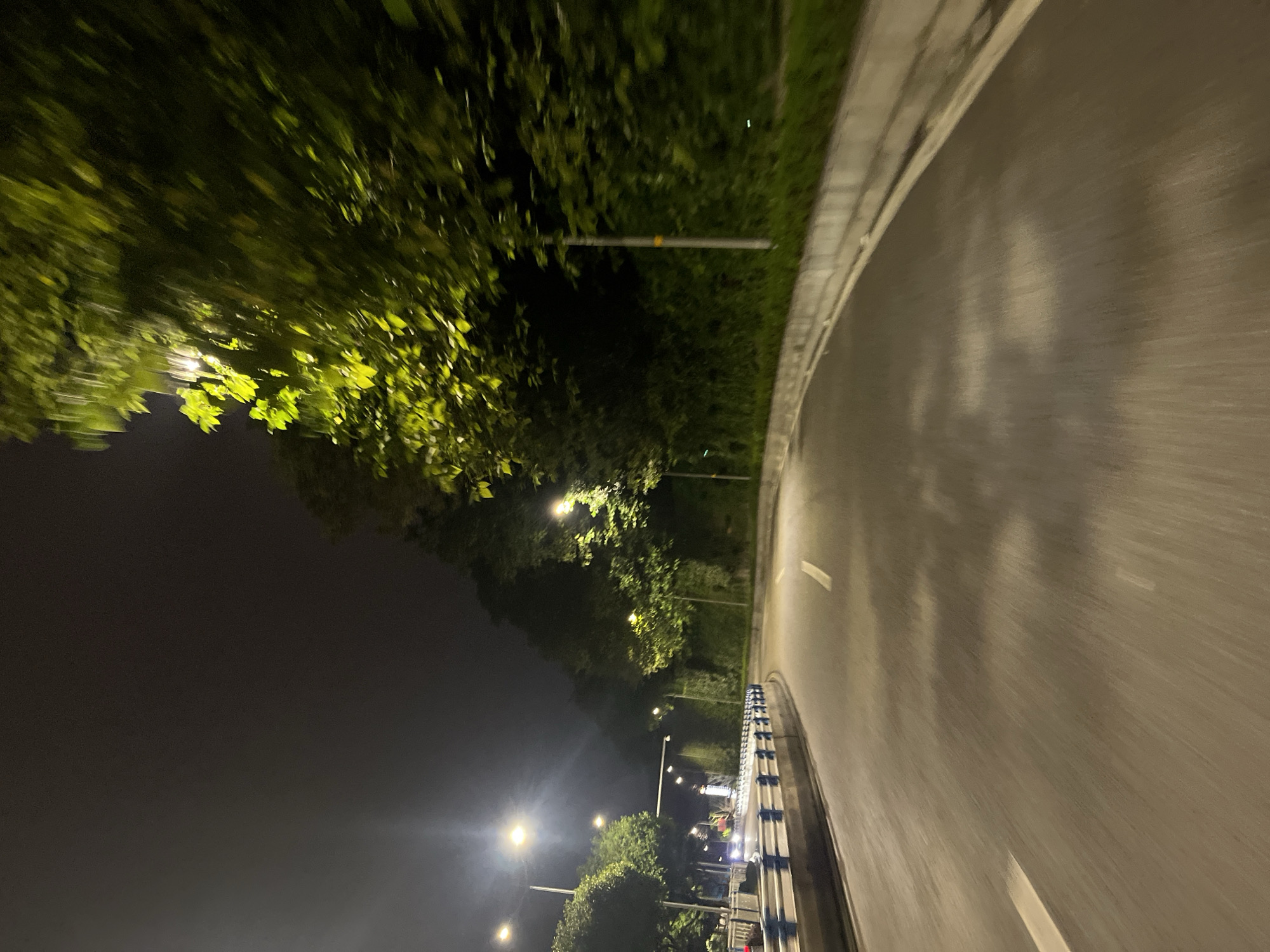 The image shows a nighttime view of a curved road in Jiulongpo District, China, likely taken from a moving vehicle. Streetlights illuminate the road, which has white lane markings. On the left side of the road, there is a blue and white barrier, followed by trees and more streetlights. On the right side, there are trees and a concrete barrier, also illuminated by streetlights. The overall scene depicts a typical urban road at night, with the motion blur indicating movement.