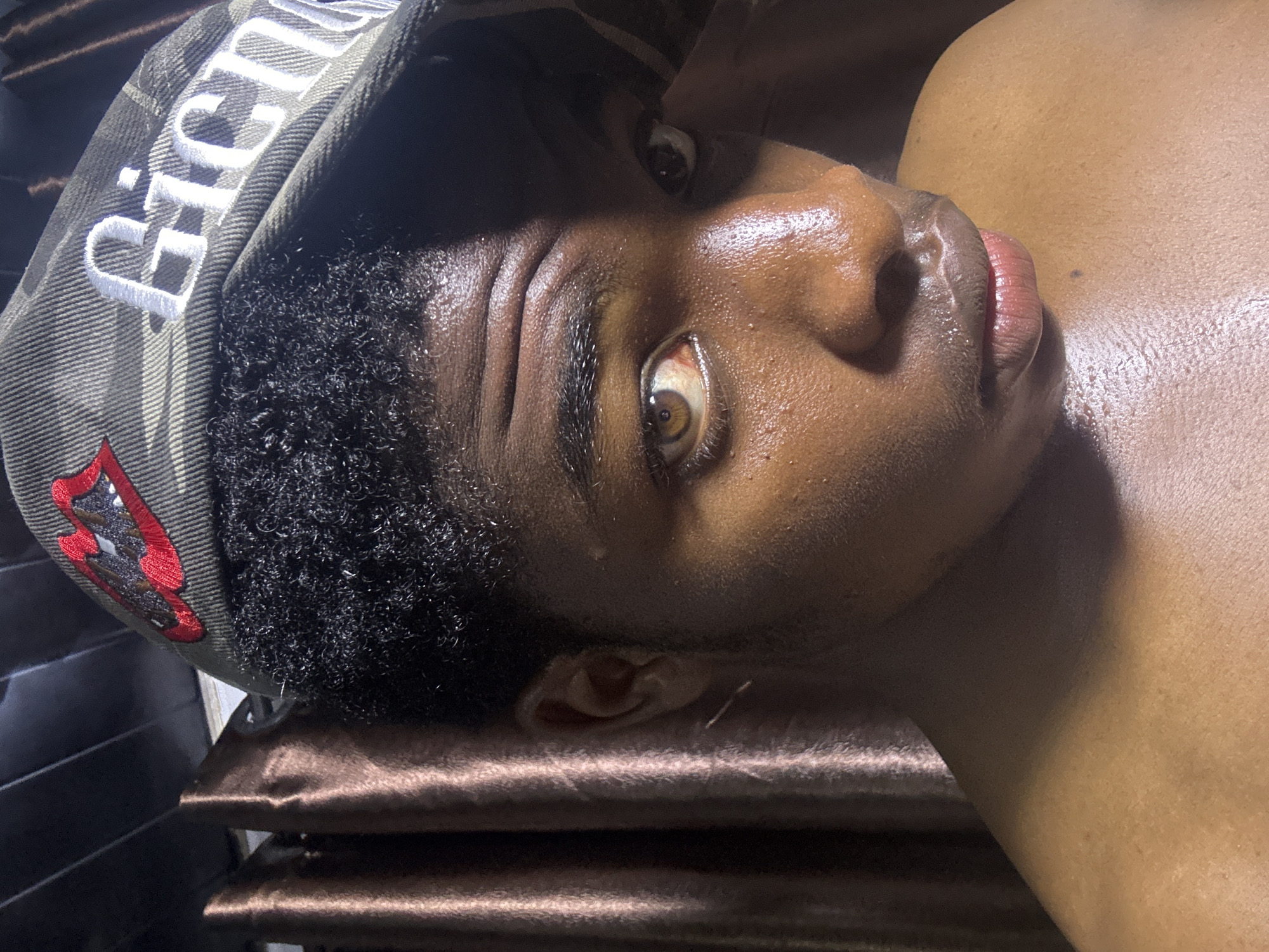 A close-up, low-angle shot shows a Black person lying on a brown pillow. The person is wearing a camouflage baseball cap with white lettering and a red lips graphic. Their skin appears to be slightly glistening. The location cannot be determined as Bukuru, Nigeria, from visual data alone.
