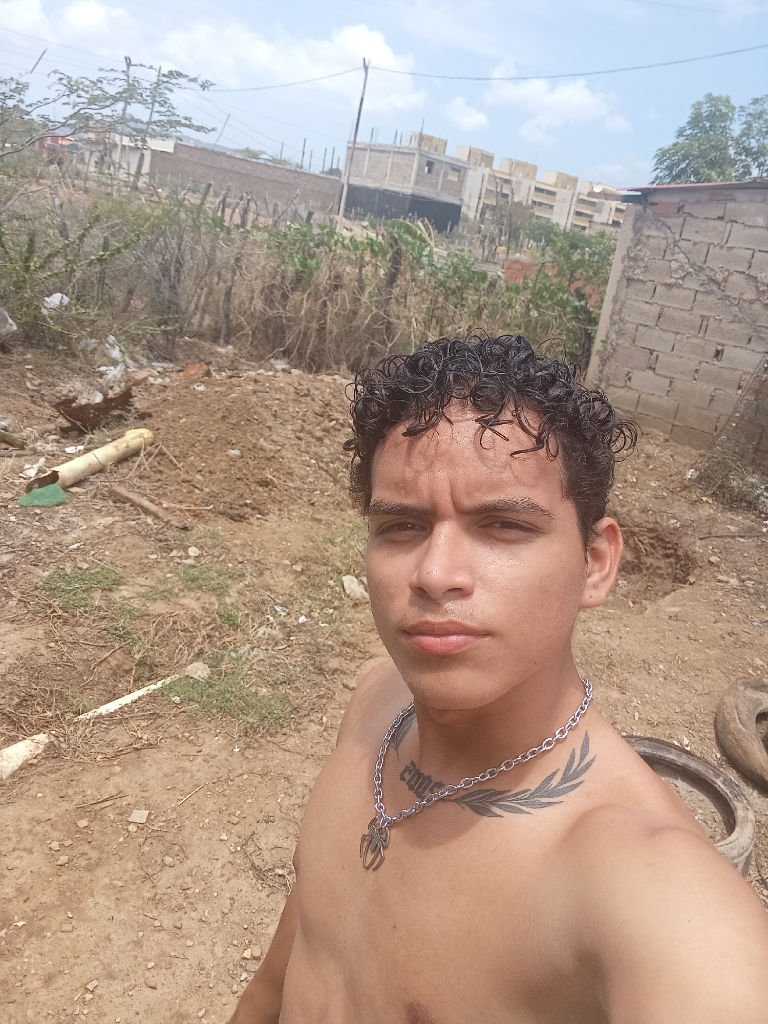 A young man with curly dark hair is standing in an outdoor area with a dusty ground. He is shirtless and wearing a silver chain with a spider pendant. He has tattoos on his chest and neck. In the background, there are unfinished buildings, vegetation, and some debris. The scene appears to be in a developing or construction area. The lighting suggests it is daytime. The specific location within Altagracia, Venezuela cannot be determined from the visual information alone.