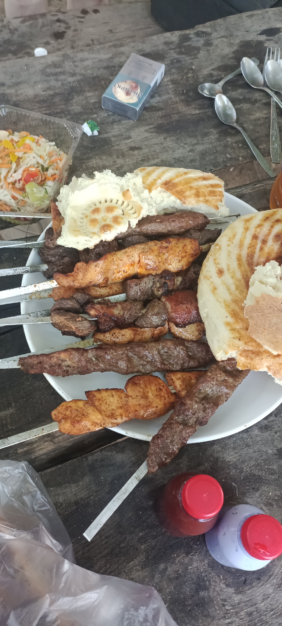 An overhead shot captures a spread of food items and assorted objects laid out on a weathered wooden table, suggesting an outdoor or rustic setting. The focal point is a large white plate filled with an assortment of grilled meats on skewers, interspersed with pieces of flatbread and a small container of salad. The meats appear to be a mix of beef and chicken, with varying degrees of char and seasoning. To the left of the plate, a clear plastic container holds a portion of coleslaw or mixed salad. A pack of cigarettes rests on the table to the upper left of the plate, next to a small piece of green wrapper. Several metal spoons and a fork are scattered around the upper right of the frame. In the lower portion of the image, two plastic bottles with red lids are visible, one containing a dark reddish liquid and the other a lighter colored liquid, possibly sauces or condiments. A plastic bag, partially obscuring the lower left corner, adds to the casual and informal presentation. The overall scene evokes a sense of a shared meal or a gathering. The image is undated and the location within "разъезд 42, Kazakhstan" is not visually identifiable from the provided image.