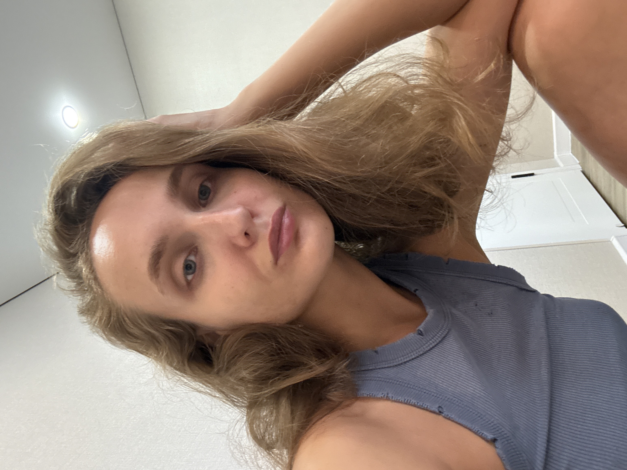 A young woman with long, wavy blonde hair is captured in a close-up selfie. She is wearing a grey ribbed tank top and has her left arm bent, with her hand resting on the back of her head. Her head is tilted, and her gaze is directed towards the camera. Her facial features are visible, including her blue eyes and slightly parted lips. The background is a plain, light-colored wall, with a ceiling light visible in the upper right corner. The setting appears to be indoors, possibly a room in a home. The image does not provide any specific geographical information to link it to the city of Подшивалово, Russia.