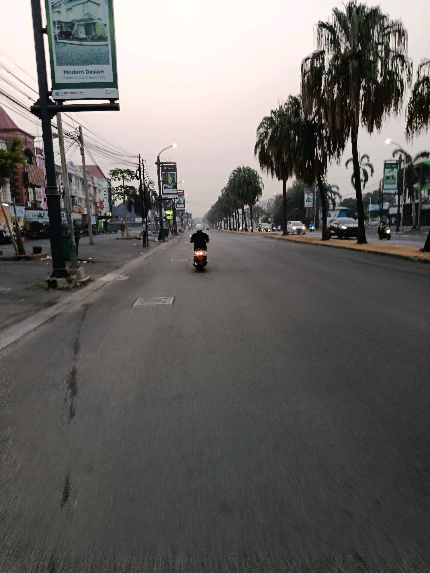 This is a low-angle, eye-level shot of a street in Bekasi, Indonesia, likely during early morning or late afternoon due to the soft, diffused light and the overcast sky. The road is paved and appears wet in places, suggesting recent rain. A person on a scooter, wearing a white helmet and dark clothing, is riding away from the camera down the center lane. The scooter has its brake light illuminated.
On the left side of the street, there are multi-story buildings with storefronts and balconies. A tall lamppost with a banner is prominently featured in the foreground. The banner displays an image of a modern house with the text "Modern Design Meets Natural Harmony" and a phone number. Further down the street, there are more buildings and trees.
On the right side of the street, a row of tall palm trees lines the sidewalk. Beyond the trees, there are more buildings and traffic. Several cars and other scooters are visible in the distance, moving in the same direction as the main subject. Streetlights and traffic signals are also present along the road. The overall impression is of a moderately busy urban street in Indonesia.