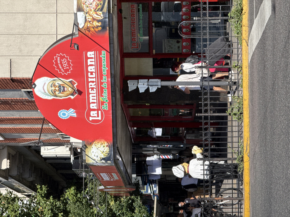 A street-level view showcases "La Americana," identified as "La Reina de las empanadas" (The Queen of the empanadas). Its red signage boasts "Calidad desde 1935" (Quality since 1935), featuring an animated chef illustration. Adjacent, the business's facade in red houses windows with "La Americana" inscribed again. Sidewalk activities are visible, including pedestrians and parked bicycles behind a metal railing near the street edge, suggesting an active urban environment.