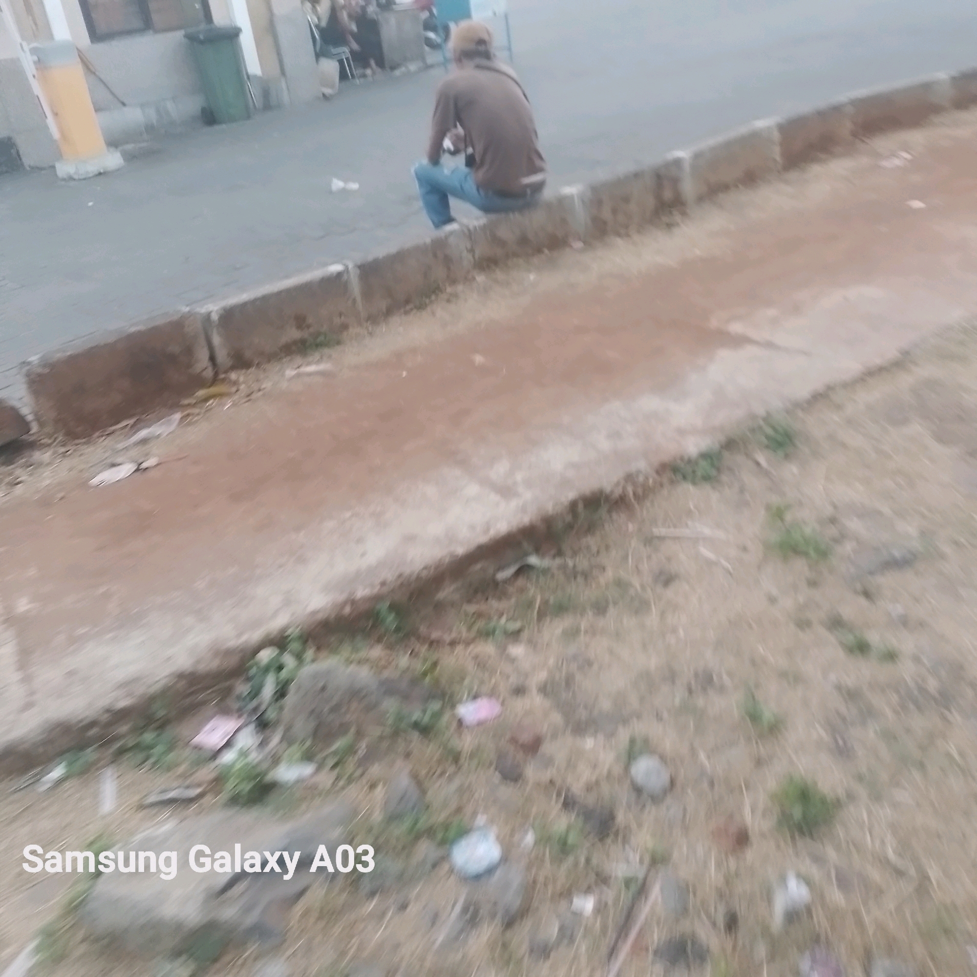 A person wearing a brown shirt and blue jeans, with a tan cap, is sitting on a concrete curb. The person's back is to the camera. The environment appears to be an outdoor urban setting. To the left of the curb, there is a paved area. Beyond that, a building with windows is visible, along with a green trash bin and a yellow pole. To the right of the curb, the ground is covered with dry grass and scattered debris. The text "Samsung Galaxy A03" is overlaid on the bottom left of the image.