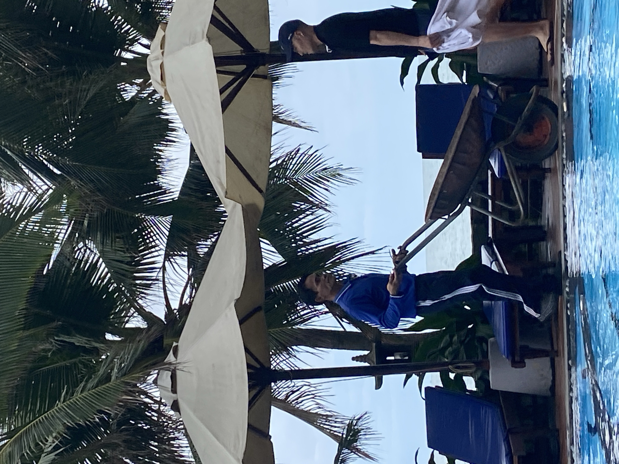 A horizontally-oriented photograph shows a tropical outdoor scene with palm trees and a swimming pool, possibly located in Phu Thuy Ward, Vietnam. On the left, the fronds of palm trees rise from the bottom edge of the frame, partially obscured by a large white parasol. To the right of the palm trees, a man wearing a blue long-sleeved shirt and dark pants sits, appearing to hold or interact with an unseen object. Behind him, there are blue chairs and lush green foliage.
Near the top of the image, another person wearing a black shirt, white shorts, and a dark cap stands at the edge of a swimming pool. A wheelbarrow sits nearby, and the blue water of the pool occupies the right side of the frame. The sky is visible in the background.