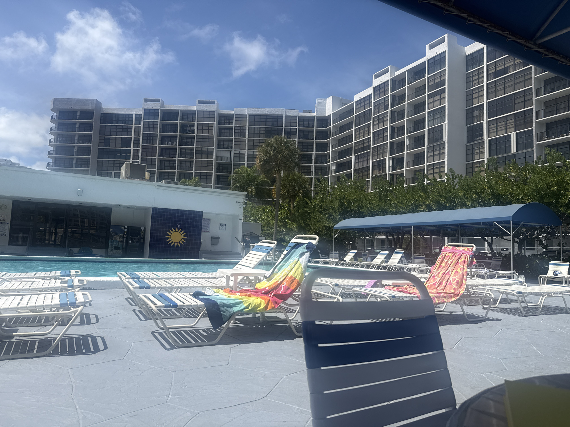 The image depicts an outdoor swimming pool area at a resort or hotel in Hallandale Beach, Florida, USA. The scene is bathed in daylight under a partly cloudy sky. In the foreground, there are several blue and white striped lounge chairs positioned on a gray-tiled pool deck. Some chairs are vacant, while others are draped with colorful beach towels. A covered seating area with a blue canopy is visible in the mid-ground. Behind the pool area, a large, multi-story building with rows of windows dominates the background. Trees and other greenery partially obscure the base of the building. To the left of the pool is a structure with a blue tiled wall bearing a yellow sun emblem. The area appears unoccupied.