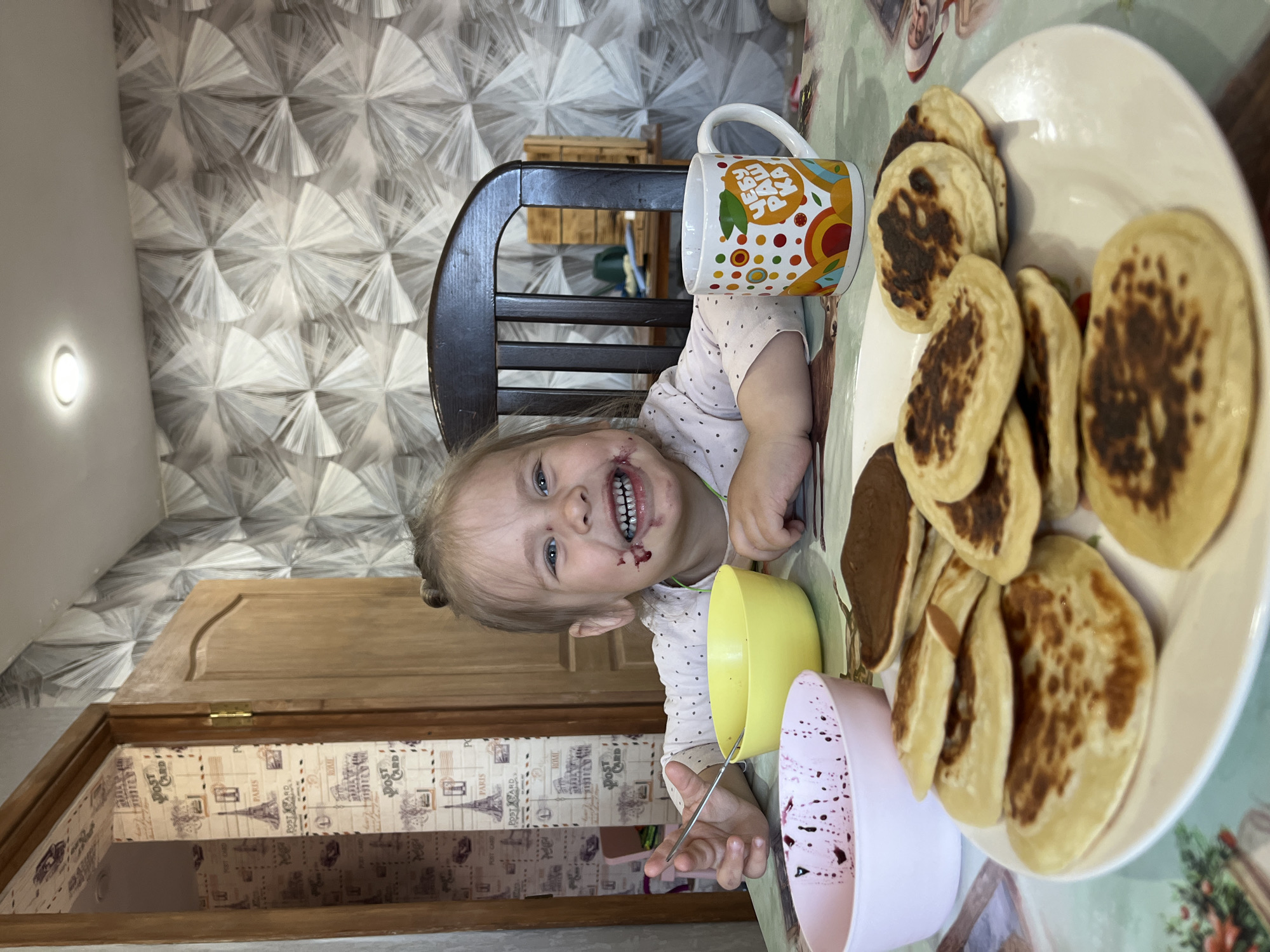 A child, likely a toddler, is seated at a table covered with a patterned tablecloth. The child has a chocolate-smeared face, indicating recent consumption of a dessert, possibly related to the contents of a yellow bowl and a pink speckled bowl, both on the table. The child is holding a spoon and smiling towards the camera. A plate of pancakes is also on the table. A mug with a colorful design sits next to the child. The room's interior shows a patterned wallpaper and a wooden door with a decorative interior, suggesting a home environment. Based on the "ЧЕБУ РАШ КА" (CHEBU RASH KA) inscription on the mug, which uses the Cyrillic alphabet, and the "PARIS" and "ROME" inscriptions on the door lining paper, the location is possibly Chaikovsky, Russia.