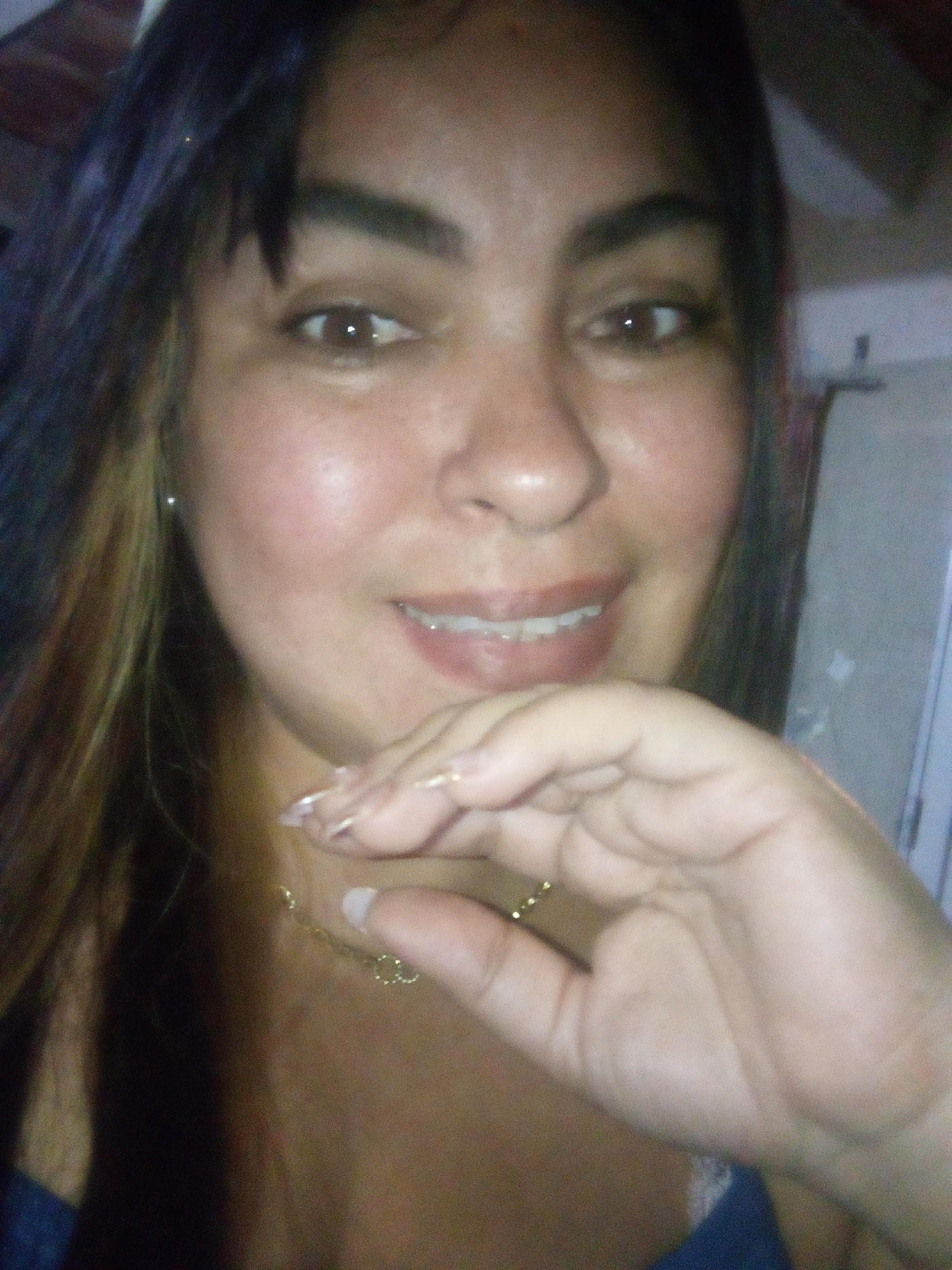 A head-and-shoulders portrait displays an adult female subject situated indoors. The subject possesses dark hair with lighter highlights, brown eyes with visible reflections, and a tan complexion. She exhibits a slight smile, with her lips parted to reveal her upper teeth. Her right hand, featuring a thin gold-colored ring on the ring finger and manicured nails with white tips, rests near her chin, partially obscuring her lower face. A delicate gold-colored chain necklace is visible around her neck, and a small silver stud earring is present in her left ear. She is attired in a dark blue garment, possibly a strap or neckline. The background is out of focus and dimly lit, revealing indistinct light-colored vertical and dark horizontal elements. The precise location within the city of Juan Griego, Venezuela, is not discernible from the visual evidence presented in the image.
