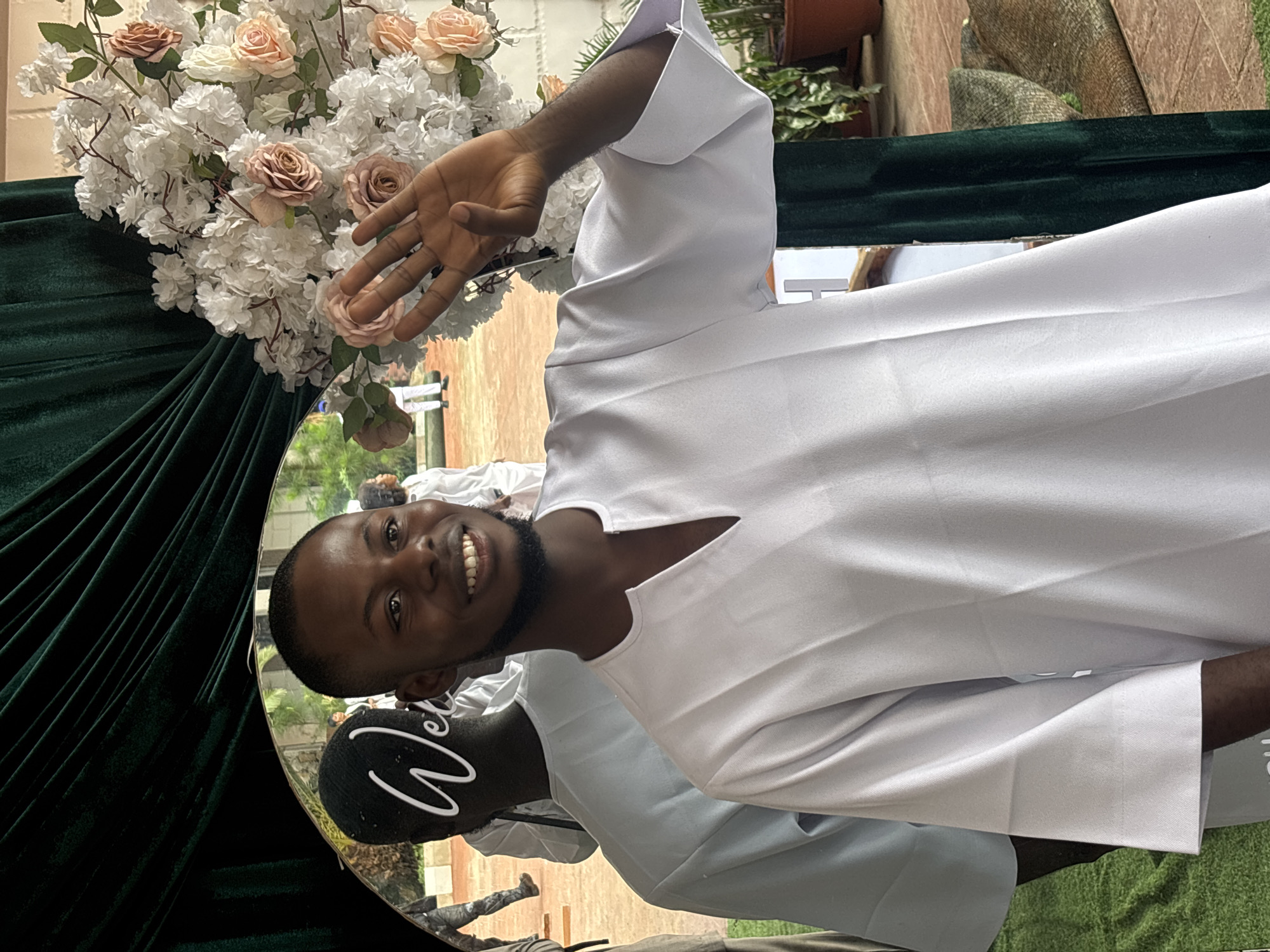 A Black male, with a beard and a beaming smile, is captured in a medium shot, looking towards the viewer and waving with his right hand. He is wearing a white, V-neck tunic. The background features a large, ornate floral arrangement of white and blush pink flowers, and a dark green velvet curtain. A mirror reflects another person in a similar white tunic, with the word "Wel" inscribed in white script. The setting appears to be an outdoor event, possibly in Jos, Nigeria, with hints of paved ground and greenery in the reflected background.