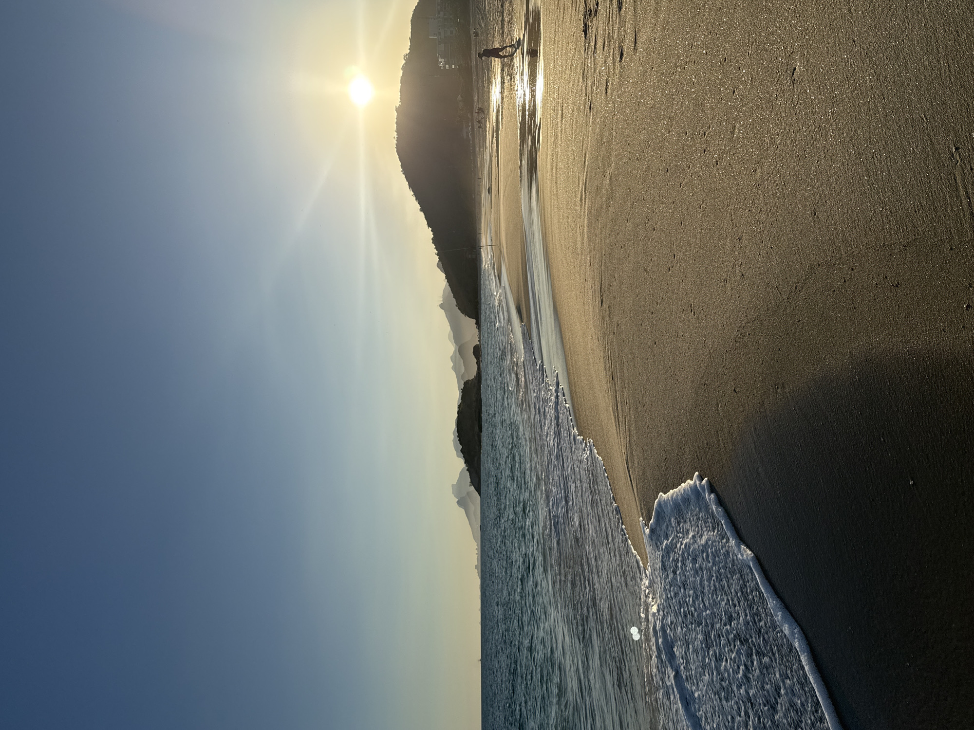 This is a vertical, eye-level shot of a beach in Niterói, Brazil, during sunset. The sun, a bright orb with visible rays, is setting behind a large, tree-covered hill in the distance, casting a warm, golden light across the scene. The sky transitions from a deep blue at the top to a lighter, hazy blue near the horizon. In the foreground, the sandy beach is wet and reflective, with gentle waves lapping at the shore. A line of white foam marks the edge of the water.
Further down the beach, silhouetted figures can be seen, including one person walking in the shallow water. Other indistinct figures and buildings are visible on the slope of the hill. The background features a chain of mountains or hills extending across the horizon, some of which are shrouded in a light haze. The overall impression is one of peacefulness and natural beauty at the close of day.