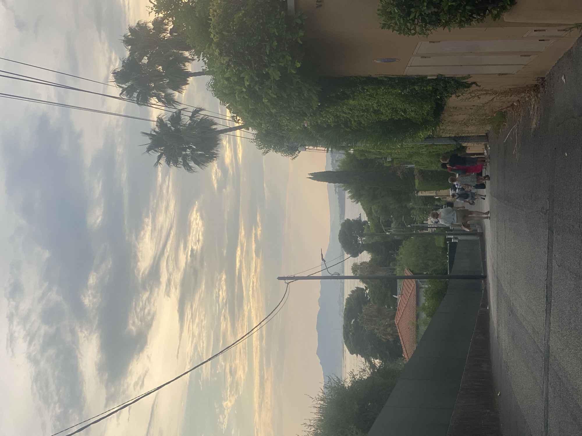 The image depicts a downward sloping asphalt road in Antibes, France, lined with lush vegetation and a light-colored building on the right. A group of people, including adults and children, are walking away from the camera down the road. The sky above is cloudy with streaks of warm light, suggesting either sunrise or sunset. In the background, a body of water is visible, with distant hills or mountains on the horizon. Utility poles and wires traverse the scene. The building on the right has a textured facade and visible panels. The overall atmosphere is tranquil and picturesque.