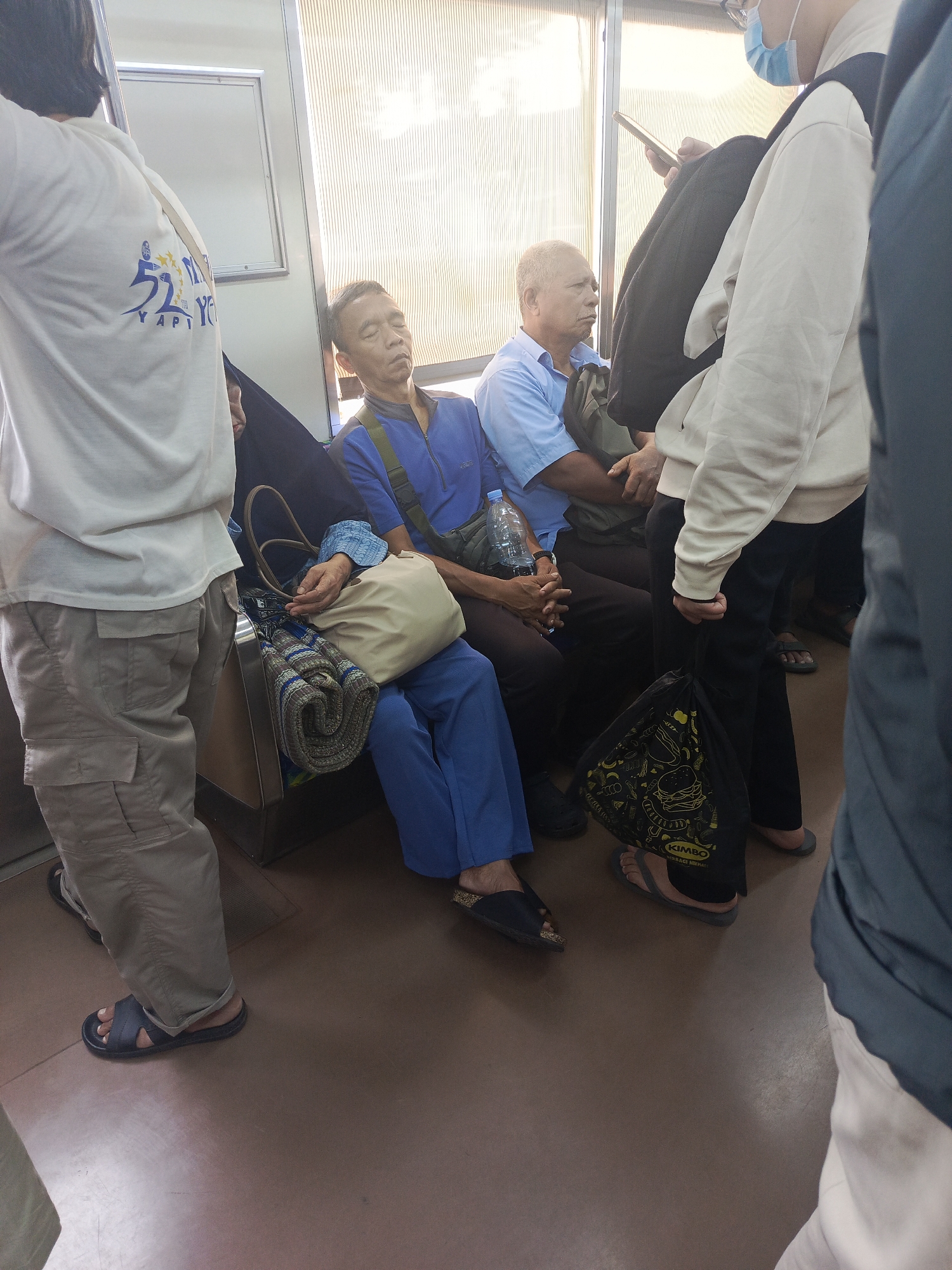 The image depicts an interior scene on what appears to be a train or bus. Several individuals are seated, with one man asleep wearing a blue shirt and blue pants, a green bag slung across his chest. Next to him, another man in a blue shirt is seated. In the foreground, a person wearing a white t-shirt with blue text and light-colored cargo pants is standing and facing away from the camera. A person in the background wears a light-colored hoodie and black pants, holding a phone. Another person is partially visible wearing a face mask. Various bags, including a beige tote bag and a black drawstring bag with food-related graphics, are present. The environment includes seating and windows with blinds. The scene is indoors, suggesting a public transportation setting within the city of East Jakarta, Indonesia.