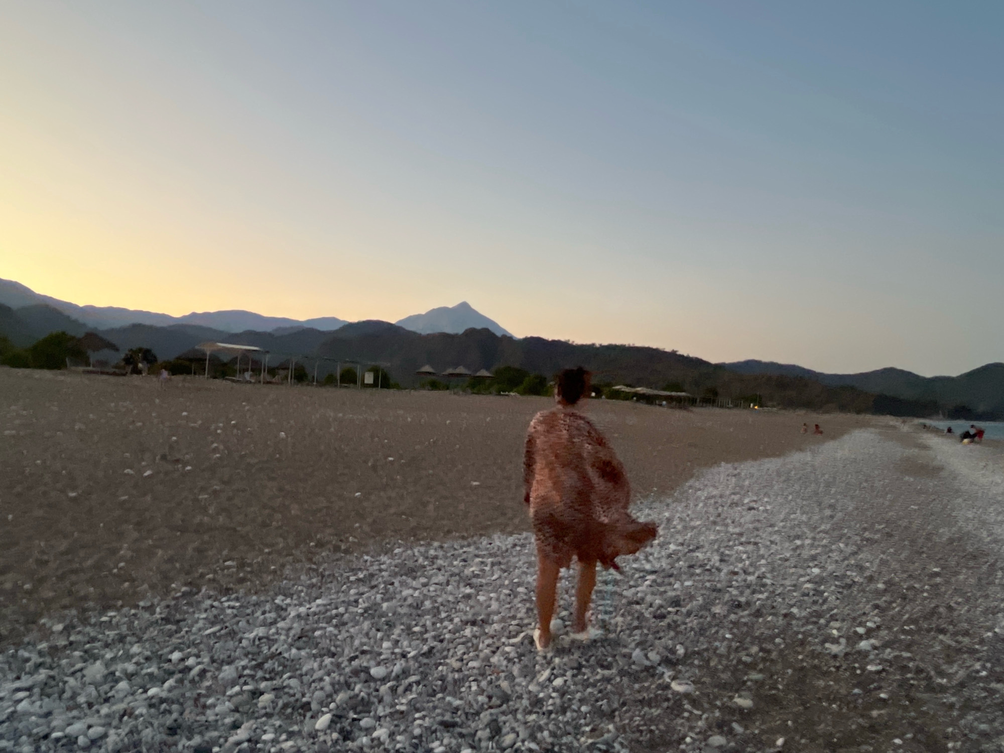 The image depicts a serene beach scene in Kemer, Turkey, during what appears to be twilight or early evening. In the foreground, a person is walking away from the camera, wearing a patterned shawl that is billowing in the breeze. The ground is covered in a mix of sand and pebbles. In the mid-ground, there is a stretch of sandy beach with a few scattered umbrellas and structures, suggesting a resort or public beach area. Further in the background, a range of hills or mountains forms a natural backdrop against a pale, clear sky that transitions from a warm yellow near the horizon to a soft blue. A prominent, lone mountain peak is visible in the distance. The overall atmosphere is tranquil and natural.