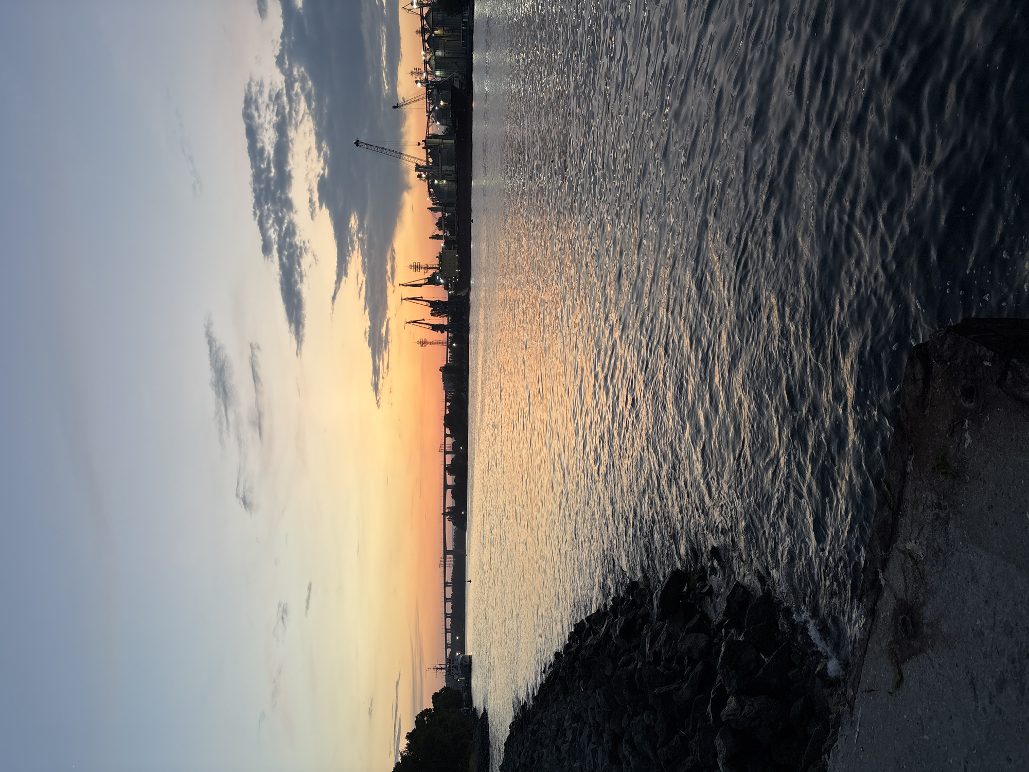 The image depicts a panoramic view of an industrial waterfront at sunset or dusk.
The upper portion of the frame is dominated by the sky, which transitions from a clear, soft blue on the left to warm orange and yellow hues near the horizon on the right, illuminated by the setting sun. Scattered clouds, some dark and dense, others lighter and wispy, are visible across the sky.
Along the horizon line, the silhouette of an industrial port stretches across the mid-ground. Numerous cranes of varying sizes stand tall, accompanied by large industrial buildings, possibly grain elevators or warehouses. Many small, bright lights punctuate the structures, indicating ongoing activity or evening illumination. Further in the distance on the left, a bridge with multiple supports spans the water, with a small boat or buoy faintly visible near its base.
The lower half of the image is filled with water, likely a river or sea. Its surface is dark and heavily rippled, shimmering with reflections of the warm sunset light. The textured patterns of the water are clearly visible.
In the immediate foreground on the bottom right, a dark, rugged shoreline composed of rocks and concrete meets the water.
The overall scene captures the blend of industrial activity with the serene beauty of a waterfront at twilight. There is no visible text in the image.