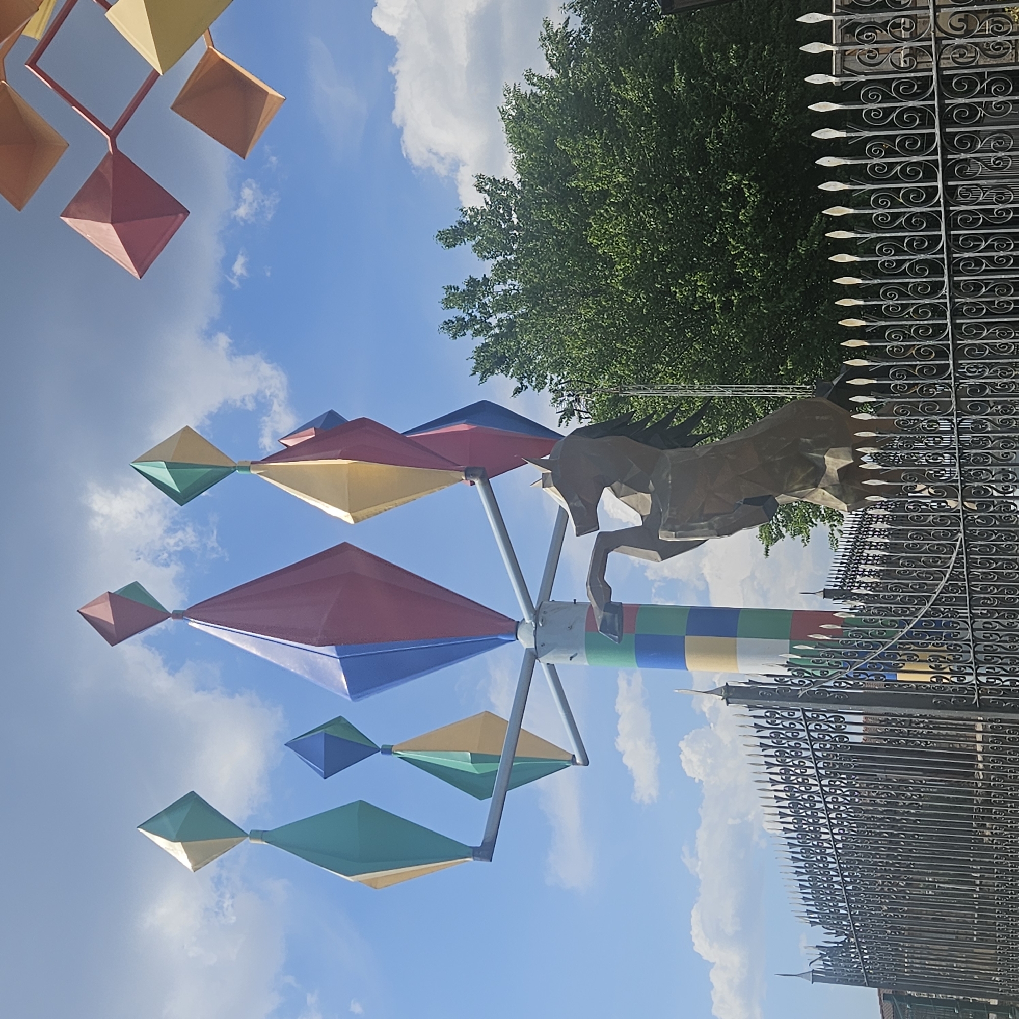 The image depicts a colorful kinetic sculpture in Algora, Spain. The sculpture features diamond-shaped wind catchers in various colors (red, blue, yellow, green) arranged around a central axis. At the top of the axis is a horse figure. Behind the sculpture is an ornate iron fence, and beyond the fence are trees and a bright blue sky with clouds. The sky is visible behind the sculpture.
