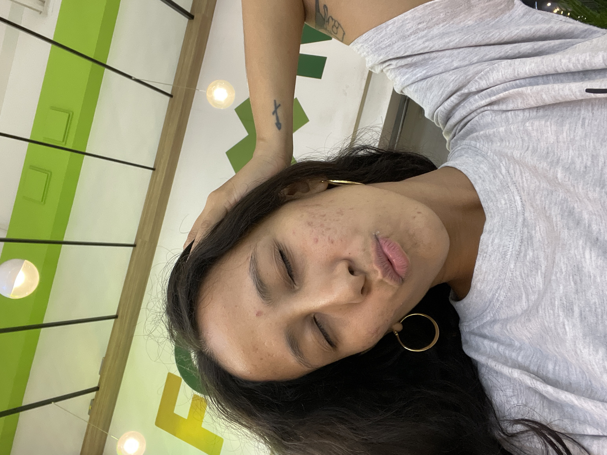 The image shows a close-up, overhead, selfie-style shot of a young woman with dark hair and closed eyes, puckering her lips. She has fair skin with visible acne scars and active blemishes on her face. She is wearing a light grey t-shirt and large gold hoop earrings. Her right arm is raised, with her hand resting on her head. A tattoo of an arrow can be seen on her inner left forearm. Behind her, a brightly lit sign with green and yellow lettering is visible, along with a white ceiling with black beams and circular light fixtures. This appears to be an indoor establishment, possibly a restaurant or cafe, given the signage and lighting. The location is identified as Porlamar, Venezuela.