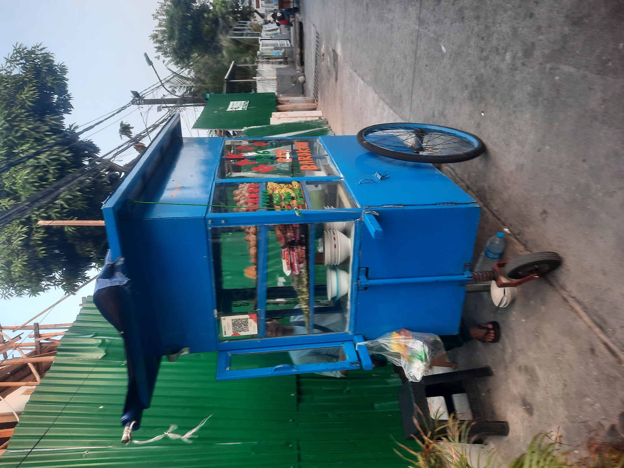 The image shows a blue food cart parked on a sidewalk next to a green corrugated metal fence, likely in North Jakarta, Indonesia. The cart has glass display cases showing various food items, including what appear to be snacks or pastries. A man is partially visible inside the cart, tending to the food. Bags of groceries are hanging from the side of the cart, and a small black stool is placed to its left. A large spoked wheel is visible on the right side of the cart. In the background, there are trees, power lines, and other structures that suggest an urban environment. The lighting indicates it is likely daytime.
