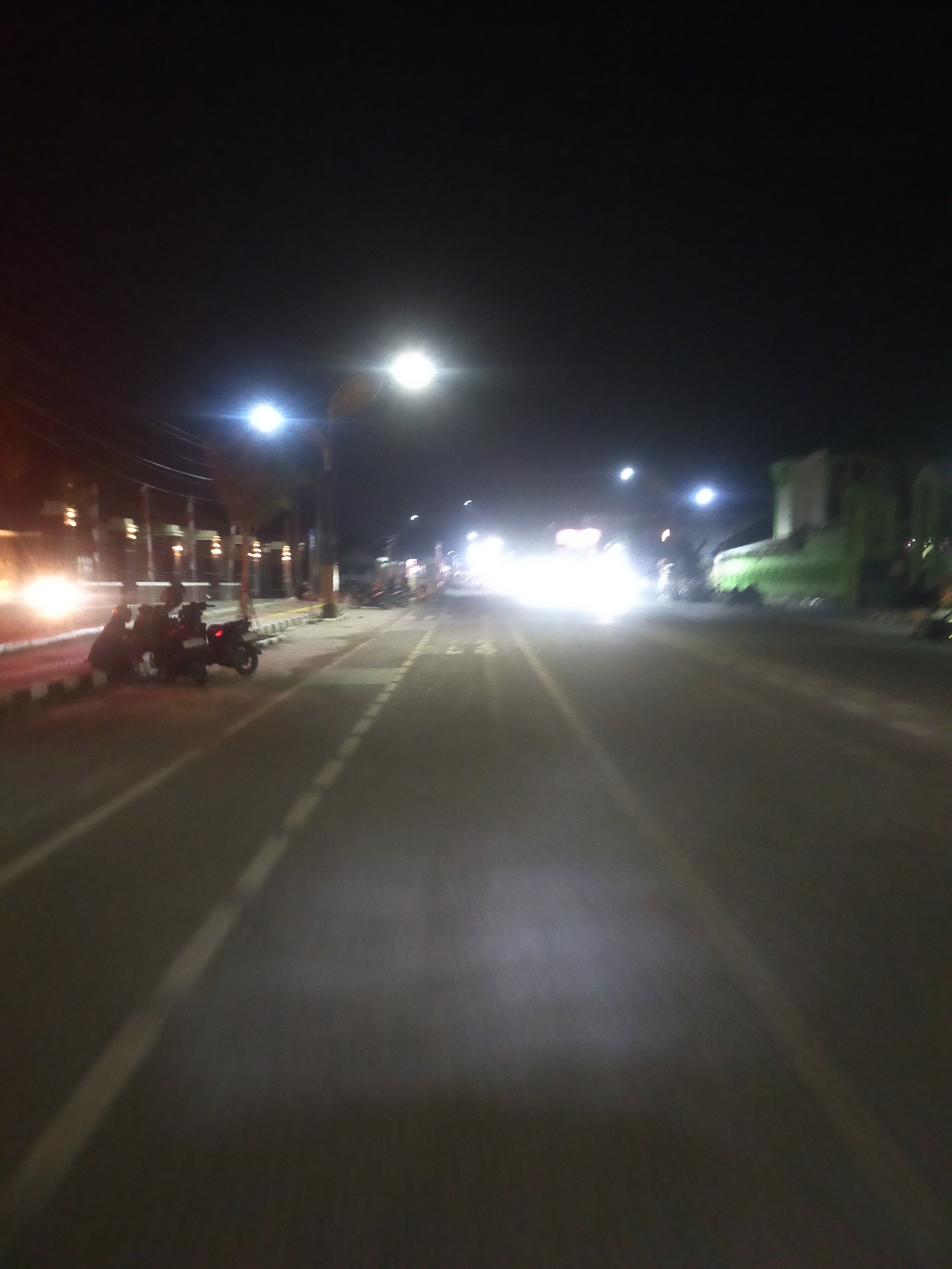 The image depicts a street scene at night in Slawi, Indonesia, characterized by low lighting and motion blur. The primary focus is a road, likely a main thoroughfare, illuminated by streetlights and the headlights of vehicles. On the left side of the road, several parked motorcycles are visible, with two individuals seated on one of them. The background includes buildings and vegetation, also dimly lit and somewhat obscured by the night and motion. The overall impression is of a busy street captured during a moment of transit, with artificial lights creating bright streaks and highlights across the scene.
