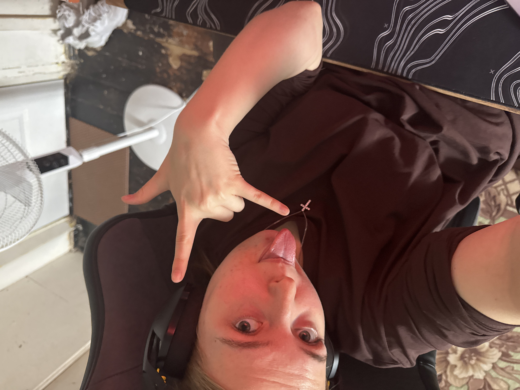 A person with fair skin and blonde hair is facing the camera, sticking their tongue out and making a hand gesture with their left hand. They are wearing a dark brown shirt and a necklace with a cross pendant. Headphones are visible on their head. The person is seated in a dark-colored chair. In the background, there is a white fan, a wooden floor, and a patterned mat. A skateboard is visible in the top right corner. The exact location in the city of Perm, Russia is not discernible from the image.