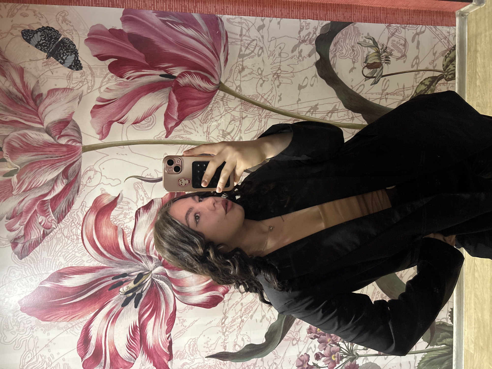 A young woman is taking a selfie. She is wearing a black top and a necklace. Her hair is dark and curly. She is holding a pink smartphone. The background is a patterned wall with large floral designs and a butterfly. The location is likely a private interior. There is no indication that this is in Perm, Russia.
