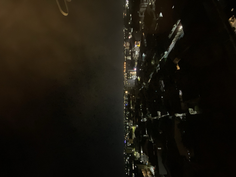 This image presents an elevated, wide-angle nighttime view of the city of Pontianak, Indonesia. The scene is dominated by a dark sky, with the urban landscape below illuminated by a myriad of artificial lights, indicating a dense residential and commercial area.
The visible environment consists of numerous low-to-mid-rise buildings, their structures largely obscured by darkness, with their presence primarily defined by illuminated windows, streetlights, and commercial signage. Linear patterns of light suggest the presence of streets and thoroughfares crisscrossing the city. A faint, diffuse glow emanates from the city lights, reflecting upwards into the lower atmosphere. No individuals, specific actions, or interactions are discernible due to the distance and low light conditions. In the upper right foreground, a distinct, light-colored, curved architectural element, possibly a railing or part of an overhead structure, partially intrudes into the frame, indicating the elevated vantage point from which the photograph was captured. The overall impression is one of a vast, sprawling urban area active under the cover of night.