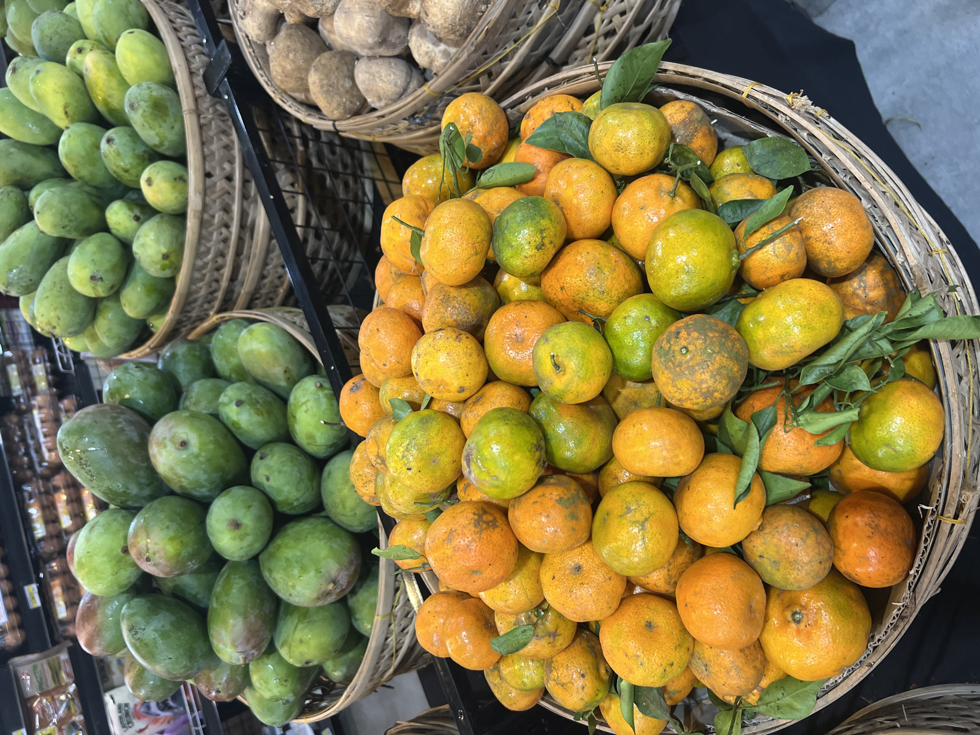 At a market stall in Sukawati, Indonesia, a close-up shot focuses on a variety of fruits displayed in woven baskets. In the foreground, a large basket overflows with ripe oranges and tangerines, their skins a mix of bright orange and green hues, some showing natural imperfections. The fruits are nestled among fresh green leaves. Behind this basket, a larger woven basket is filled with green mangoes, their surfaces smooth and varied in shade. To the right of the mangoes, another basket contains lighter-colored, spherical fruits, possibly potatoes or sweet potatoes, with rough, brown skins. The background is blurred but suggests the bustling environment of a market, with shelves and more produce visible. The lighting is natural and evenly distributed, highlighting the textures and colors of the fruits.