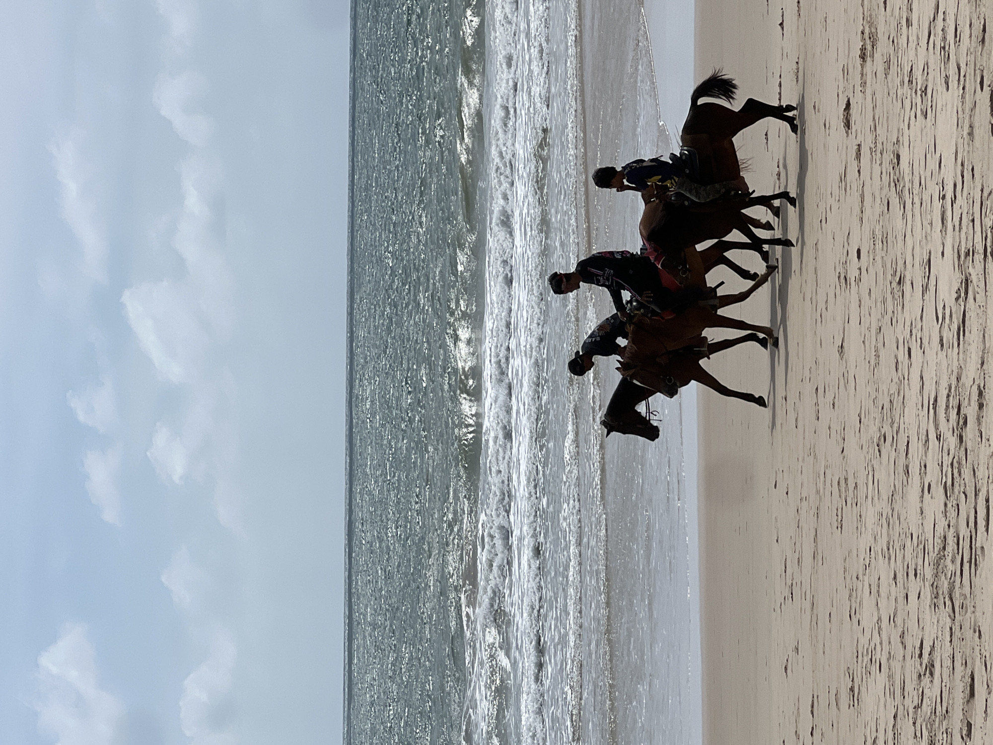 Three individuals are riding horses along the sandy shore of a beach. The sea is visible in the background with waves breaking on the shore. The sky is partly cloudy. The riders and horses are in silhouette against the brighter background of the sea and sky. One rider is wearing a bandana and dark clothing. Another rider is wearing a black shirt with a design. The third rider is wearing a blue and yellow shirt and dark pants. The horses are dark brown. The scene is likely taking place in Choeng Thale, Thailand, given the tropical appearance of the beach and ocean.
