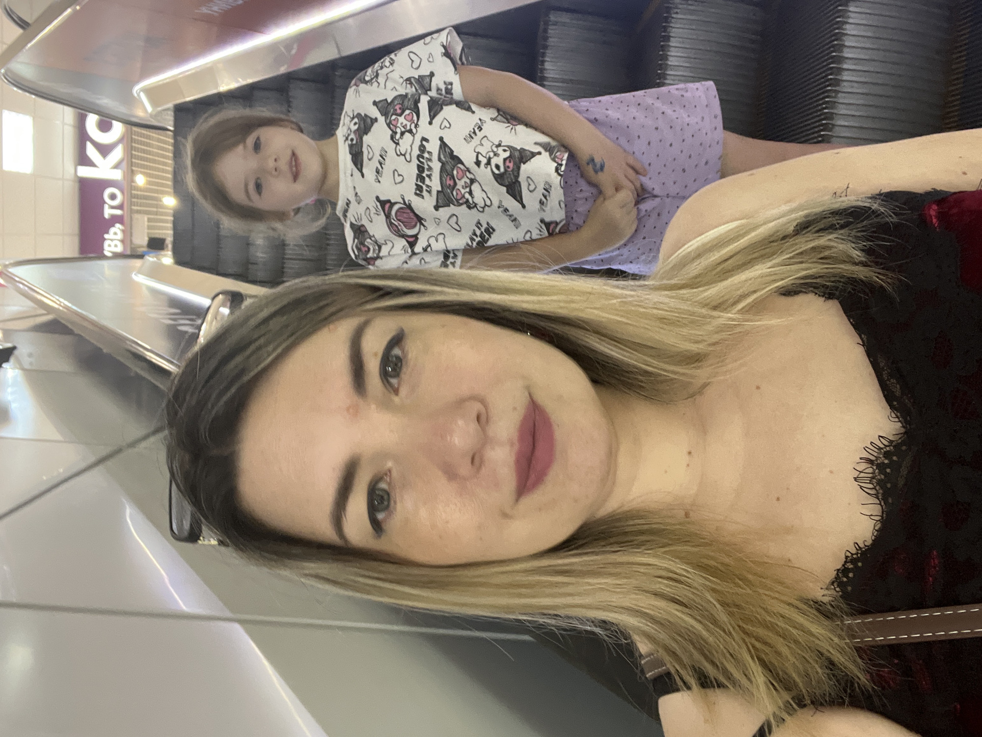 A selfie taken on an escalator features two people, a woman and a young girl. The woman is in the foreground, smiling at the camera with her blonde hair falling around her shoulders. She is wearing a black top with lace detailing. The young girl is in the background, looking towards the camera. She has blonde hair and is wearing a white t-shirt with a cartoon print and a purple skirt. The background shows the metal steps of an escalator. A sign that reads "Bb, TO KO" is visible on the left side of the image, with the word "KO" being the most legible. The setting appears to be an indoor public space, possibly a shopping mall or an airport, given the escalator and signage. The overall mood appears to be casual and happy.